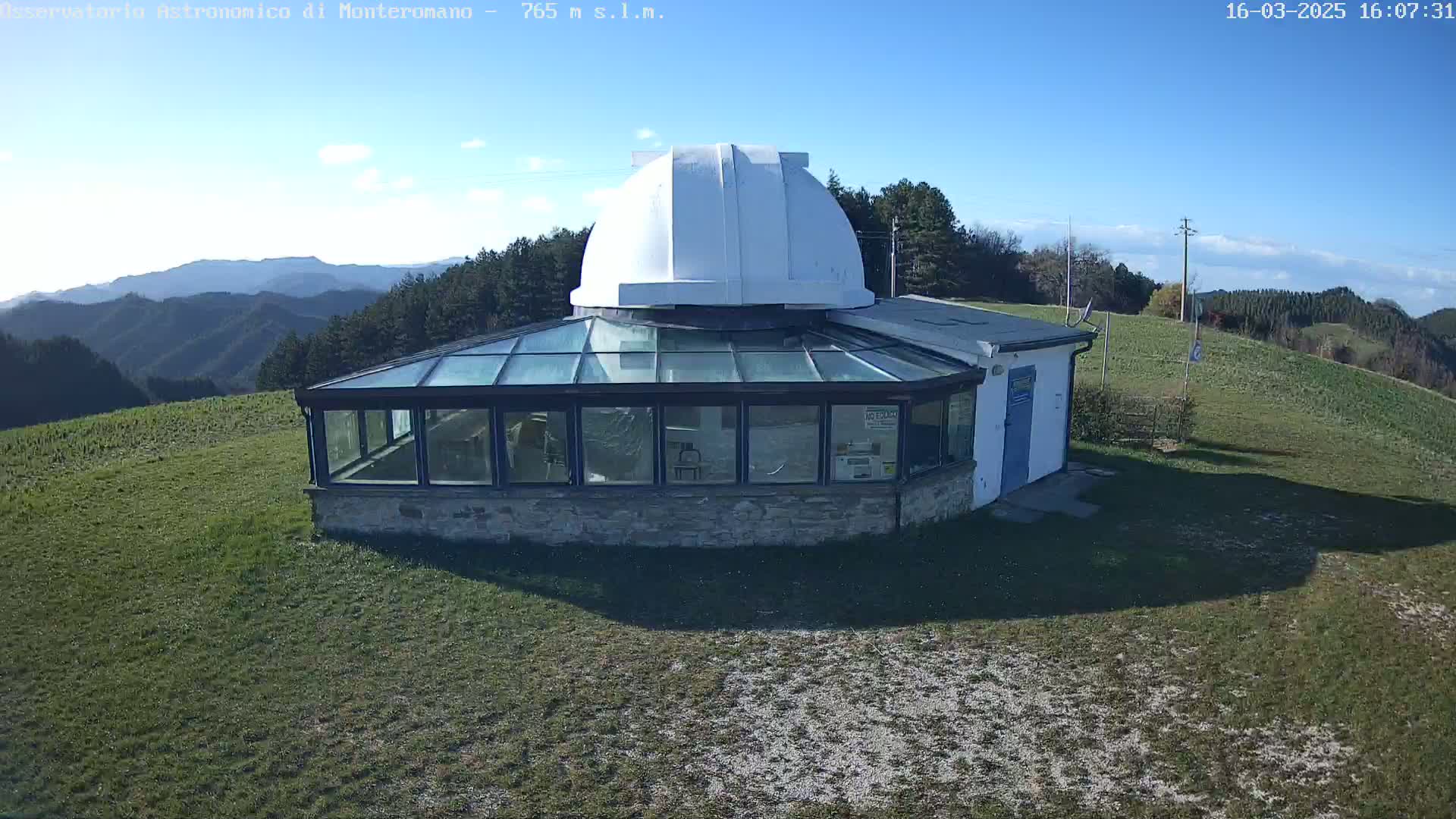 A white domed observatory building with a glass-walled addition sits on a grassy hilltop on a sunny day, overlooking a range of distant mountains.