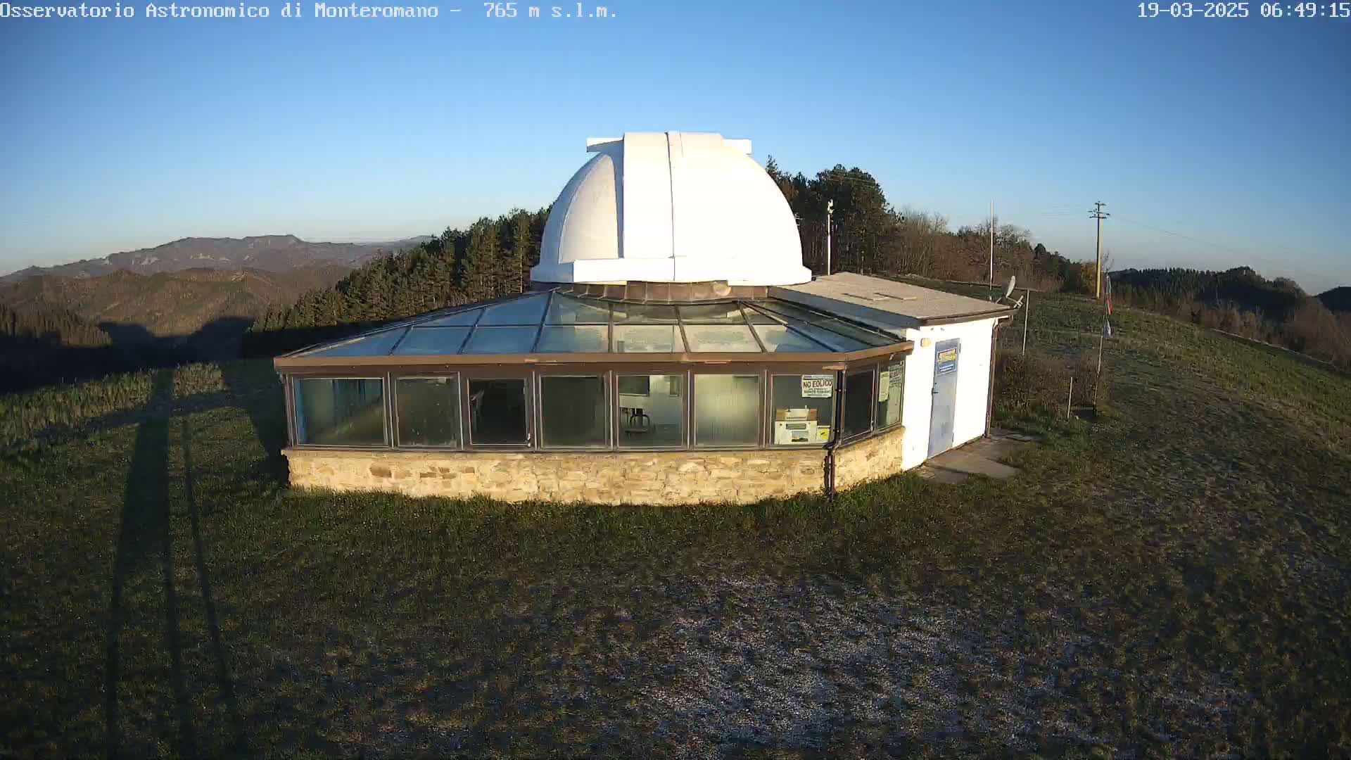 An astronomical observatory with a white dome sits atop a grassy hill under a clear blue sky.