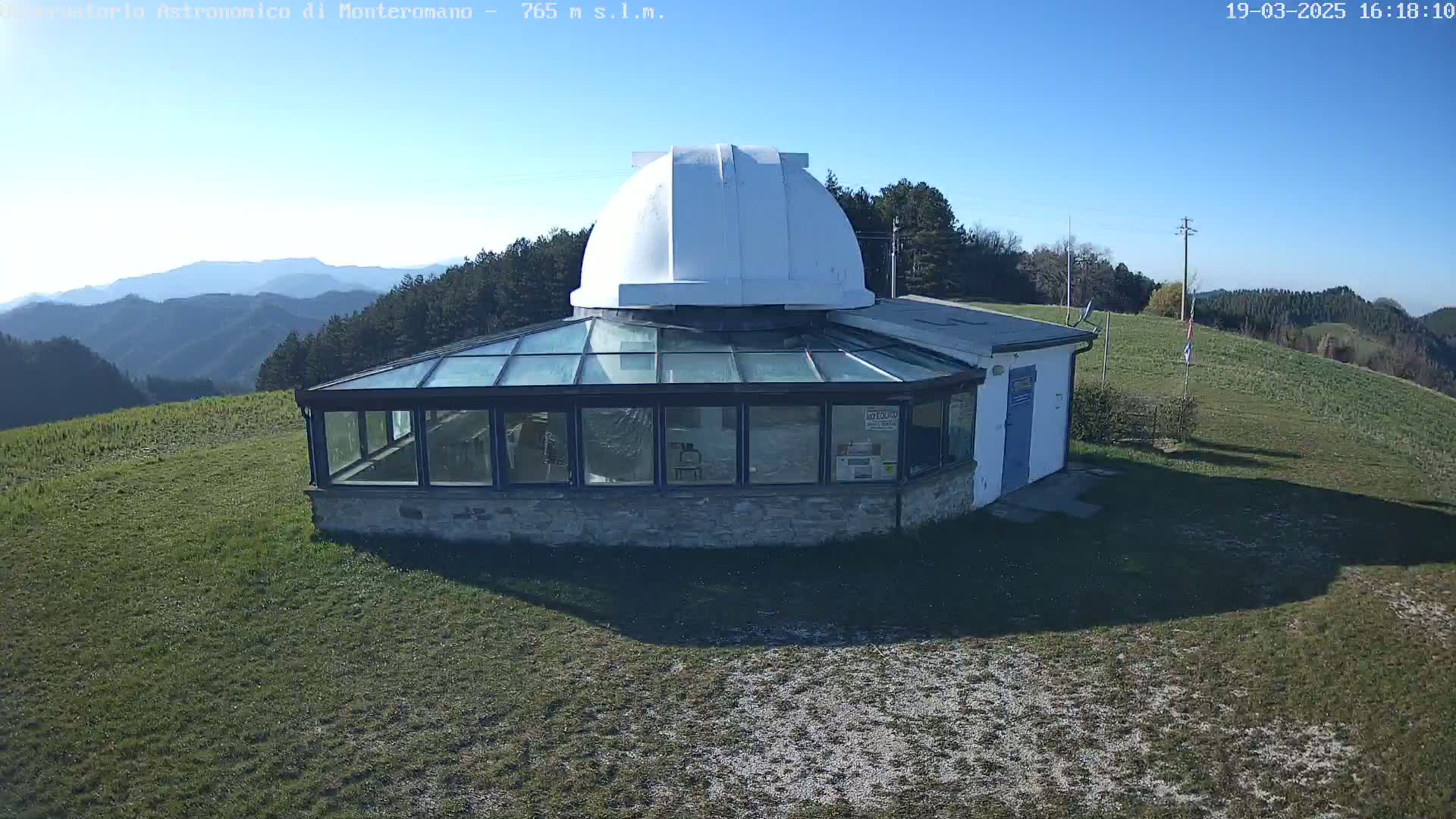 A white domed observatory building with a glass-enclosed structure is situated on a grassy hilltop under a clear blue sky.