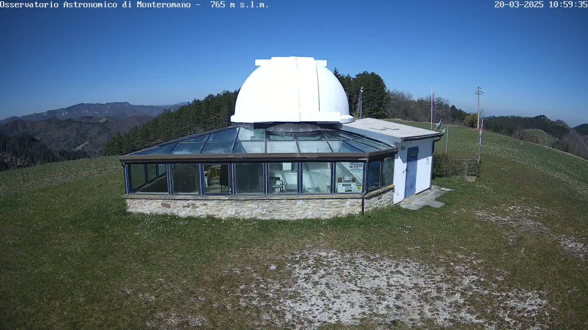 A stone building with a glass roof and a white dome on top sits on a grassy hilltop on a sunny day, with distant mountains visible in the background.