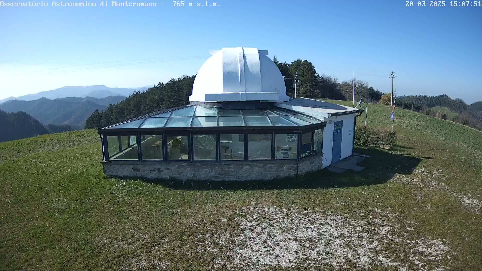 A white-domed observatory building with a glass enclosure sits on a grassy hilltop under a clear blue sky, overlooking a mountain range in the distance.
