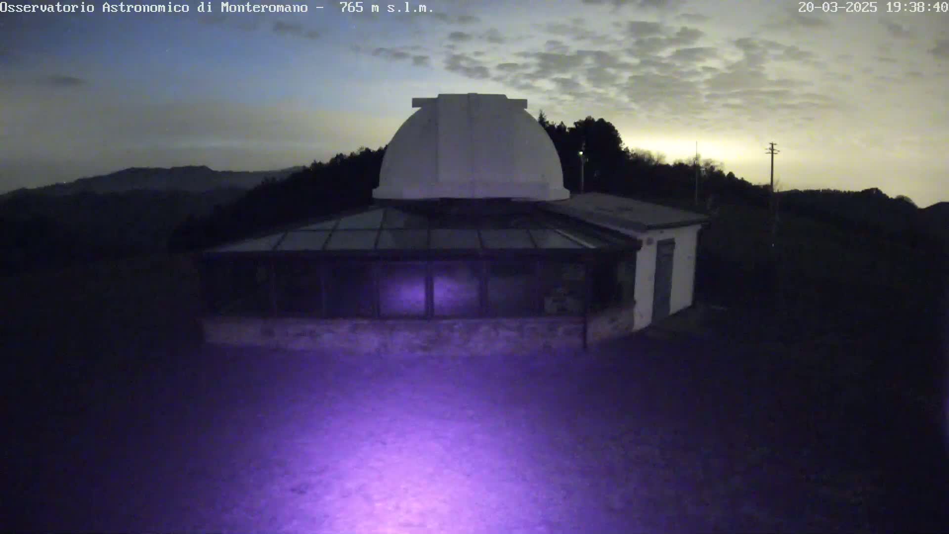 A low-light image shows a white observatory dome atop a building with a glass addition, situated on a hill under a cloudy twilight sky.