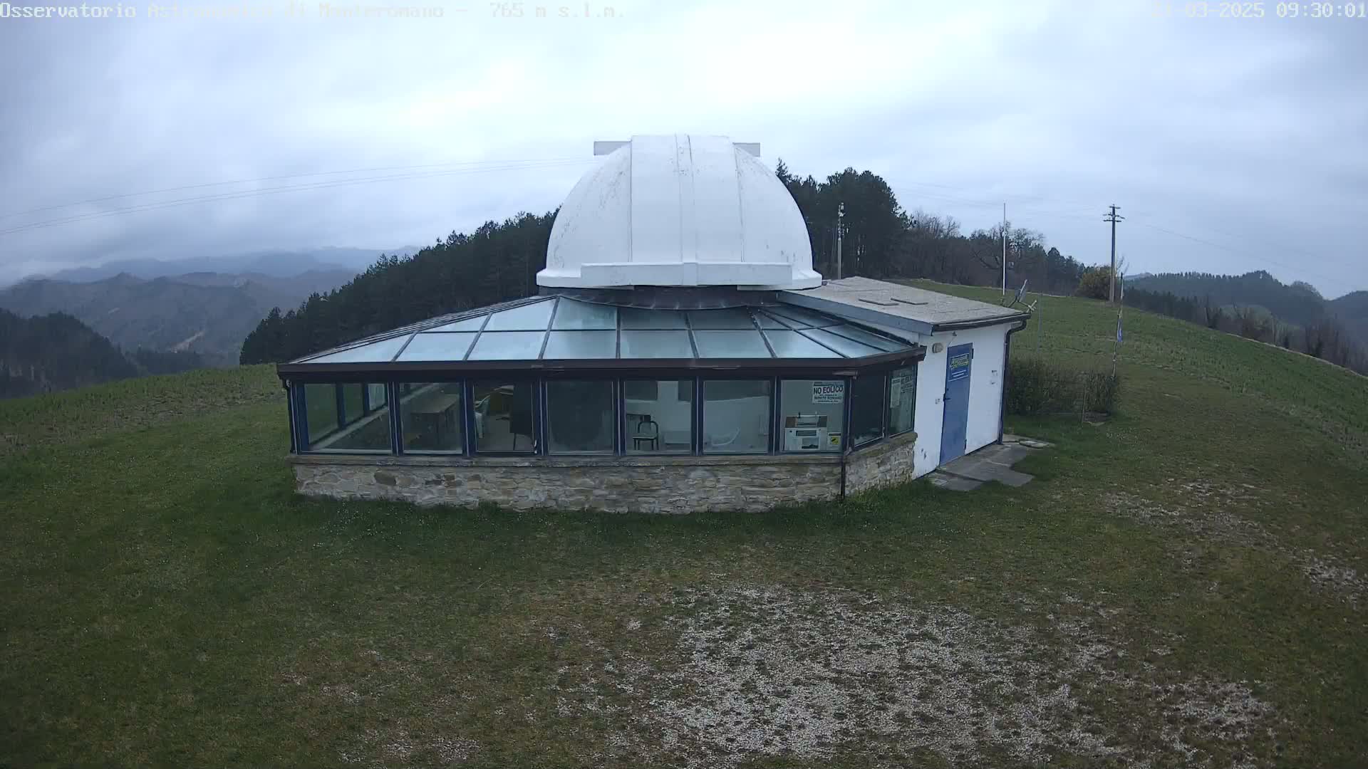 A white-domed observatory building with a glass enclosure sits atop a grassy hill overlooking a range of misty mountains under a cloudy sky.