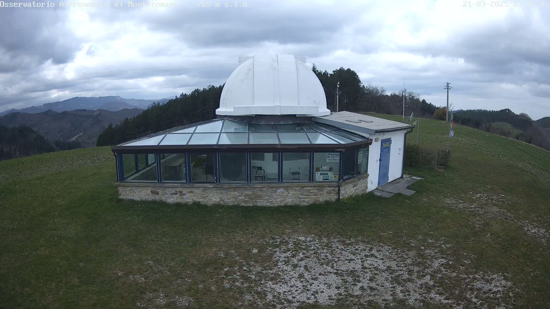 A stone observatory with a glass sunroom and a white dome sits on a grassy hilltop under a cloudy sky, overlooking a mountain range.
