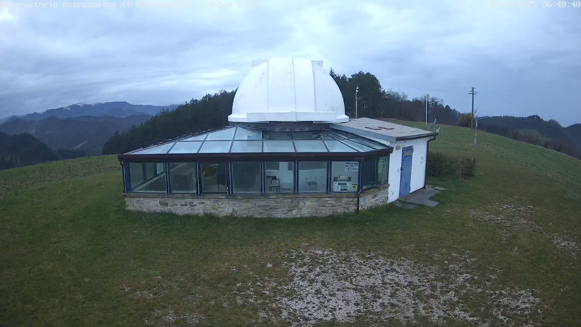 A stone building with a glass conservatory and a white dome sits on a grassy hill overlooking a range of mountains under a cloudy sky.