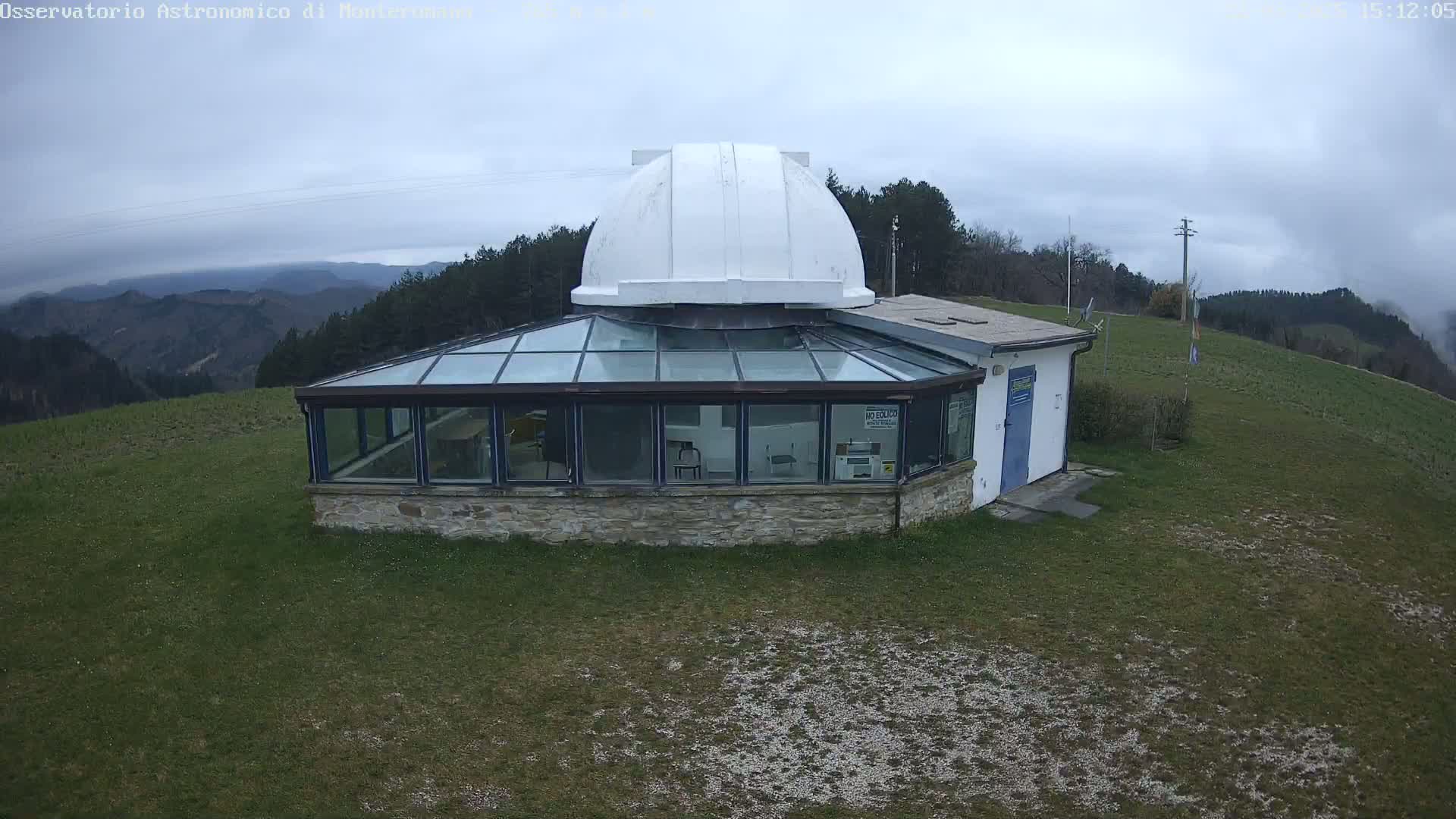 A stone building with a glass roof and a white dome sits on a grassy hilltop overlooking a hazy mountain range under an overcast sky.