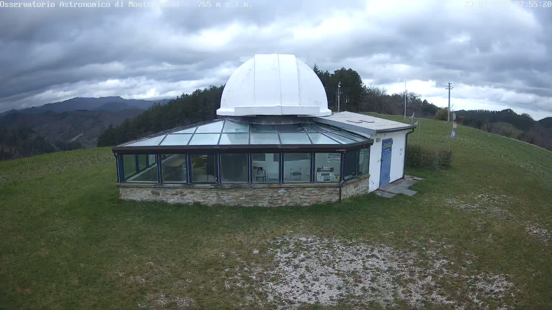 A stone building with a glass sunroom and a white dome sits on a grassy hill under a cloudy sky.