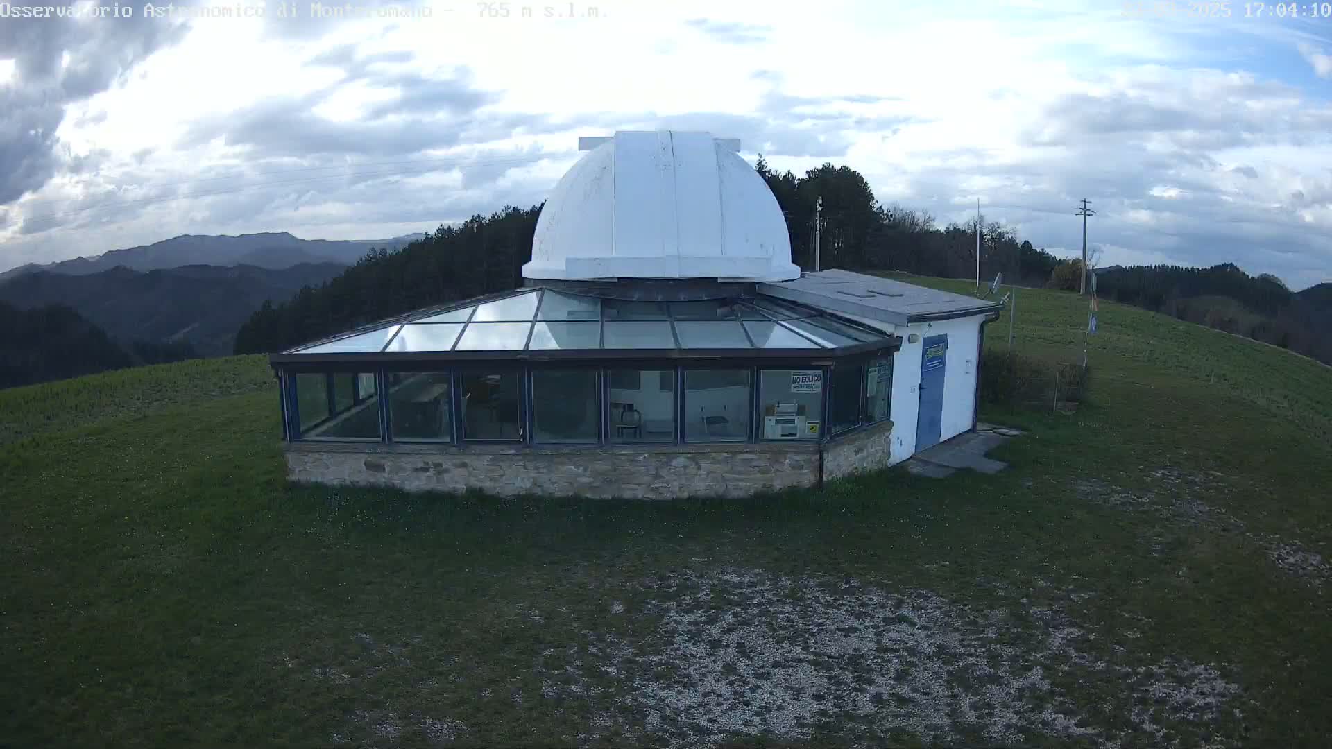 A white-domed observatory building with a glass enclosure sits on a grassy hilltop under a partly cloudy sky.