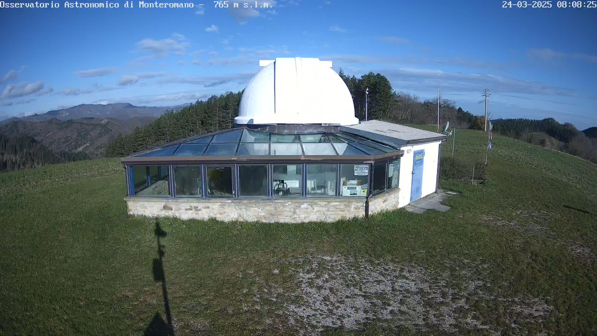 A white-domed observatory building with a glass-roofed addition sits on a grassy hilltop under a mostly clear blue sky.