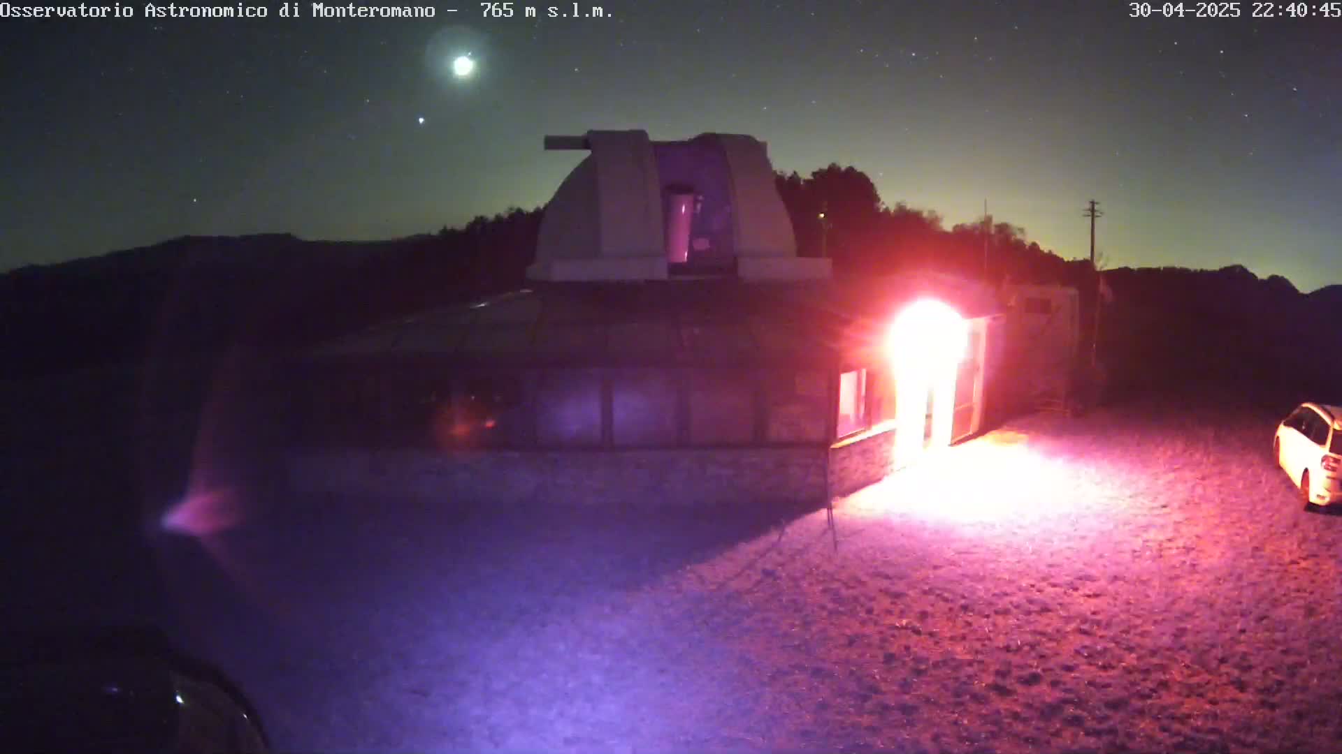 A low-light nighttime image shows a building with a dome, possibly an observatory, illuminated by a bright, reddish light emanating from its entrance, under a clear, starry sky.
