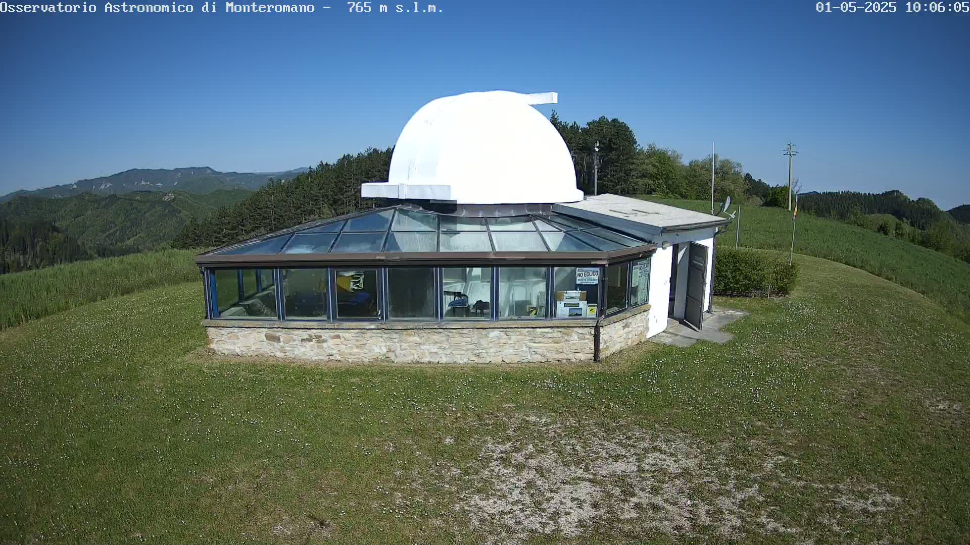 A stone building with a glass sunroom and a white dome sits on a grassy hilltop overlooking a mountain range under a clear blue sky.