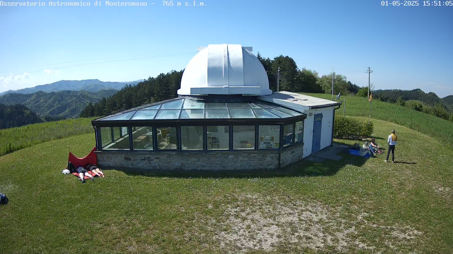A white domed observatory building with a glass sunroom is situated on a grassy hill overlooking a mountain range under a clear blue sky.