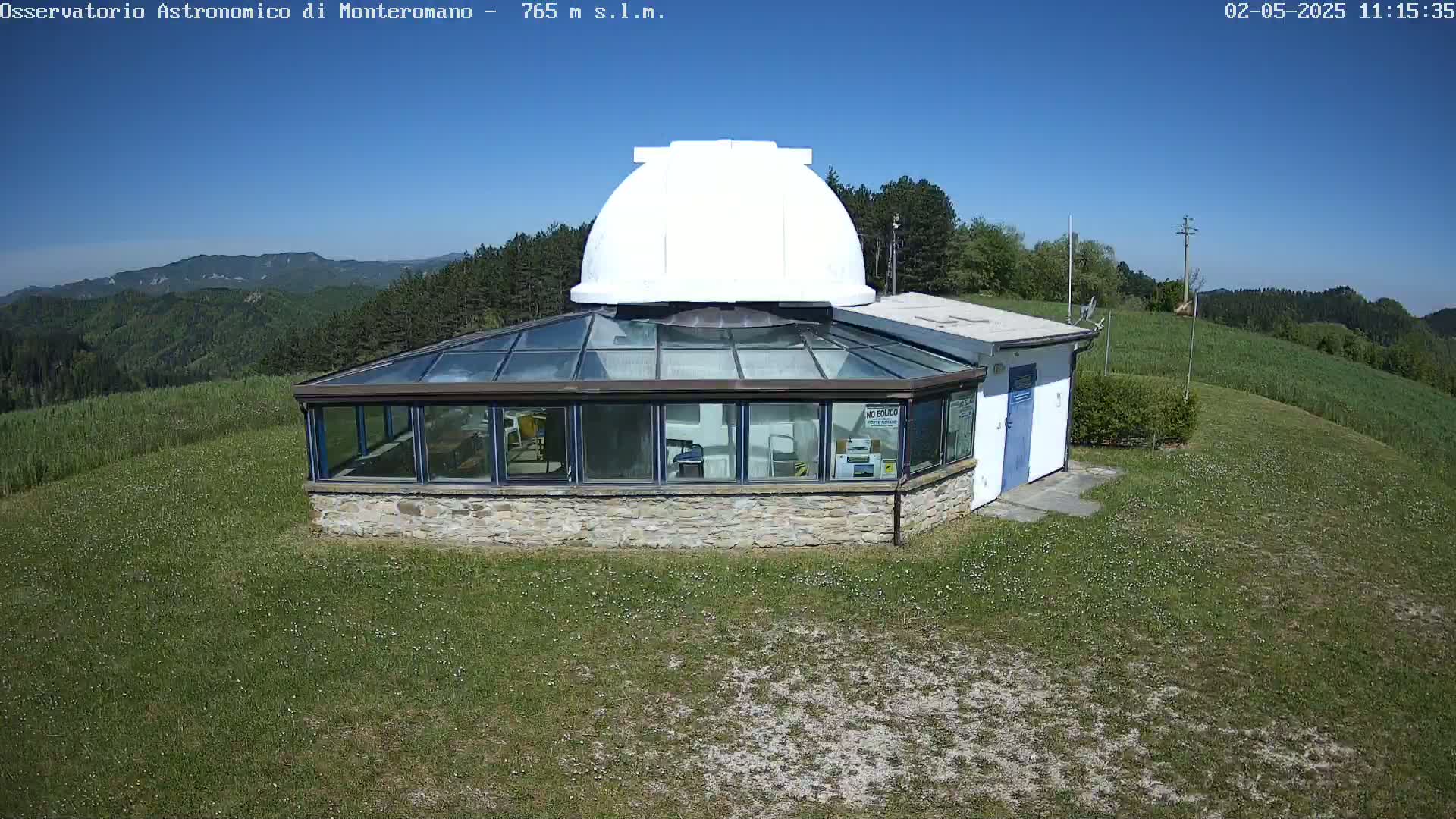 A white dome observatory sits atop a stone building with a glass-roofed addition, all situated on a grassy hill overlooking a mountainous landscape under a clear blue sky.