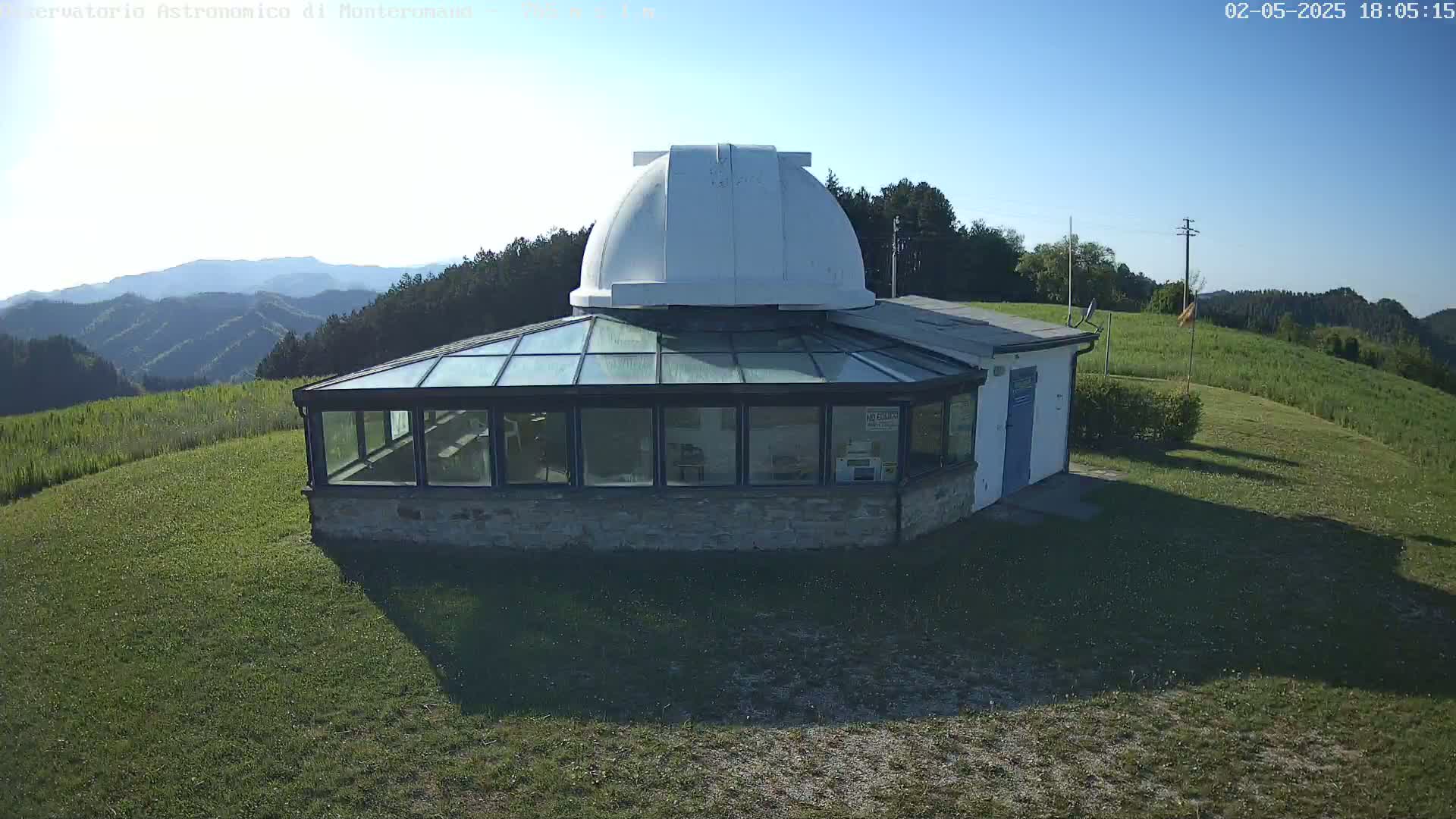 A small observatory with a white dome sits atop a grassy hill overlooking a distant mountain range under a clear, sunny sky.