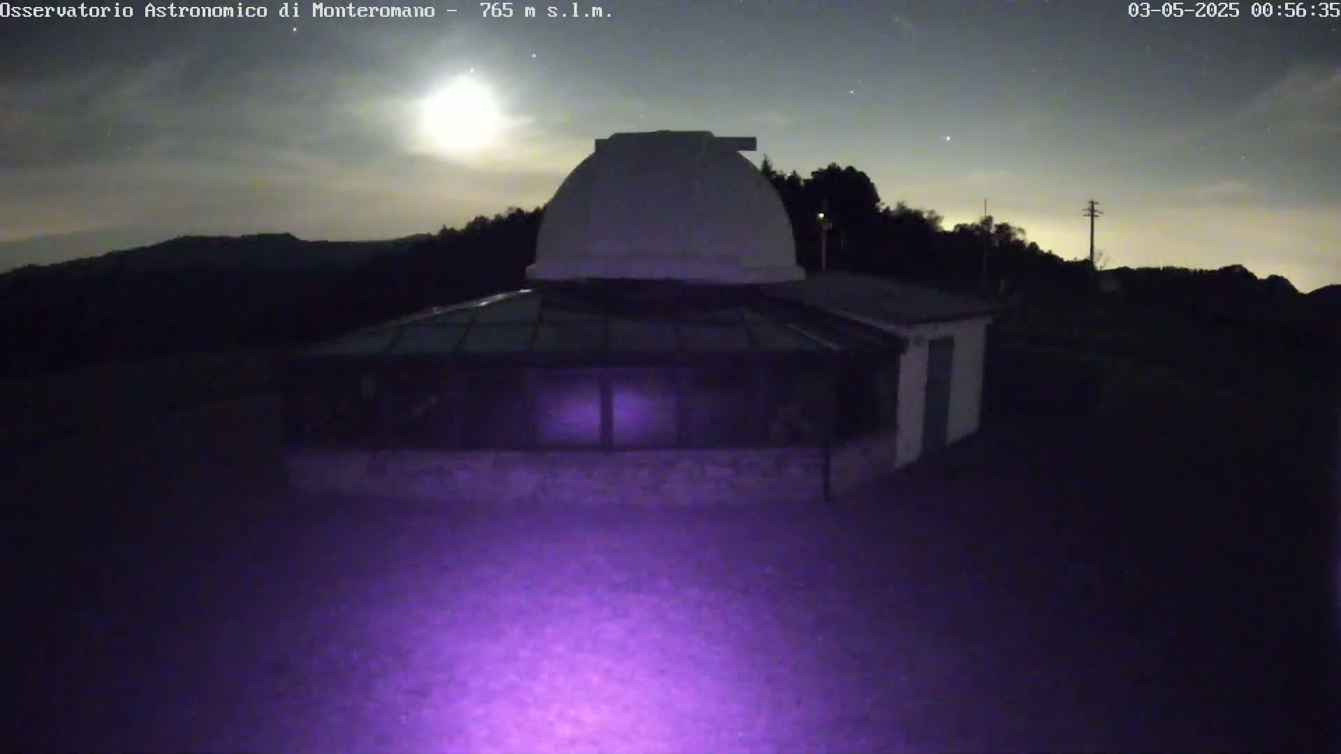 Under a mostly clear night sky with a bright moon, a low-lying building with a dome-shaped roof and glass-covered sections sits atop a hill.
