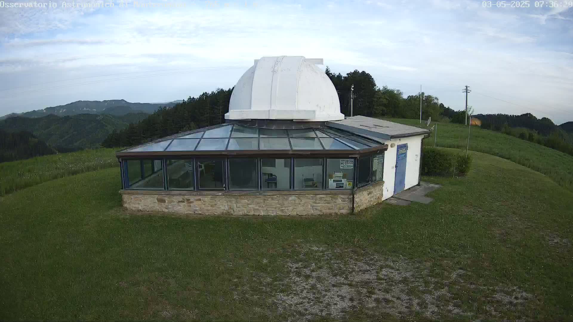 A stone building with a glass enclosure and a white dome sits on a grassy hill overlooking a mountainous landscape under a mostly clear sky.