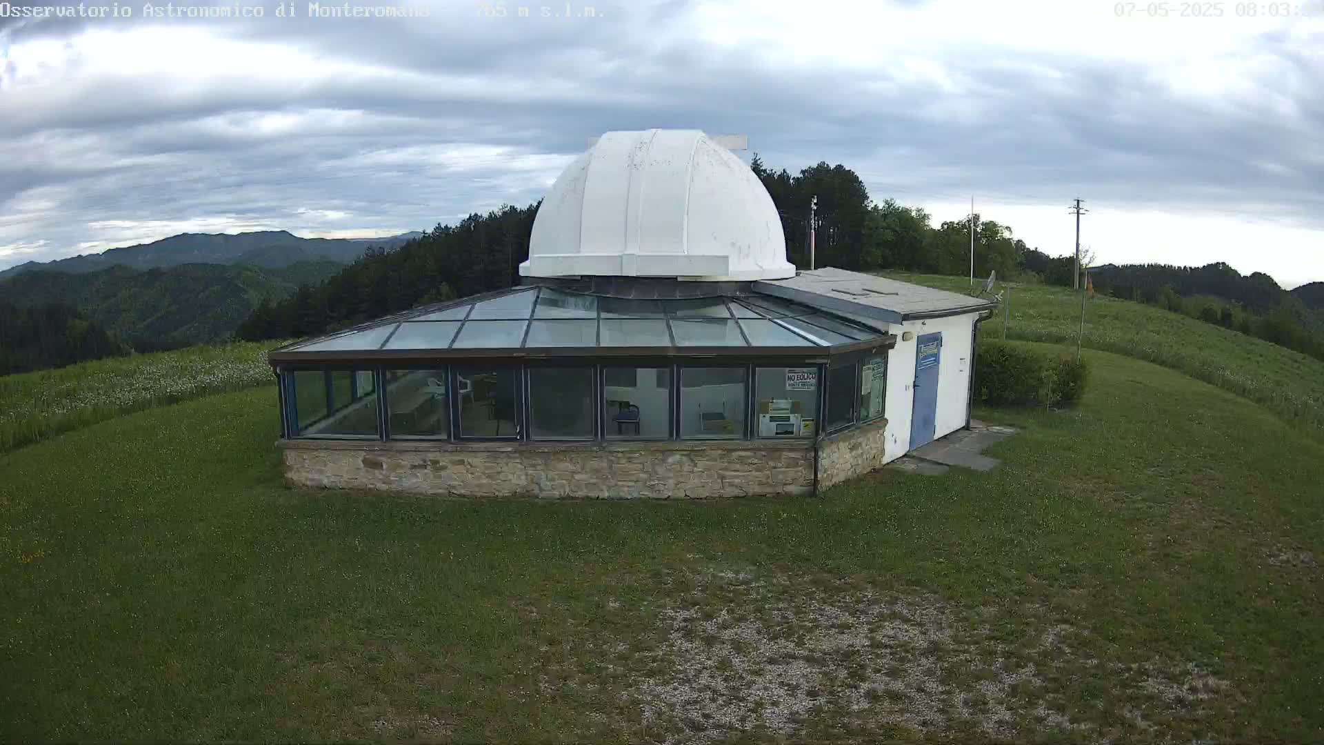 A stone building with a glass-roofed addition and a white dome sits on a grassy hill overlooking a mountain range under a cloudy sky.