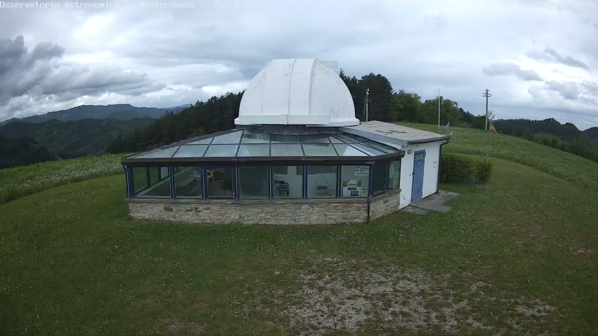 A stone observatory building with a glass enclosure and a white dome sits on a grassy hilltop under a cloudy sky.