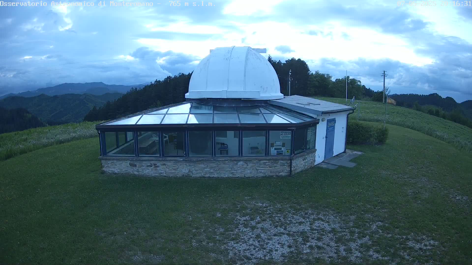 A stone building with a glass roof and a white dome on top sits on a grassy hill overlooking a mountainous landscape under a partly cloudy sky.