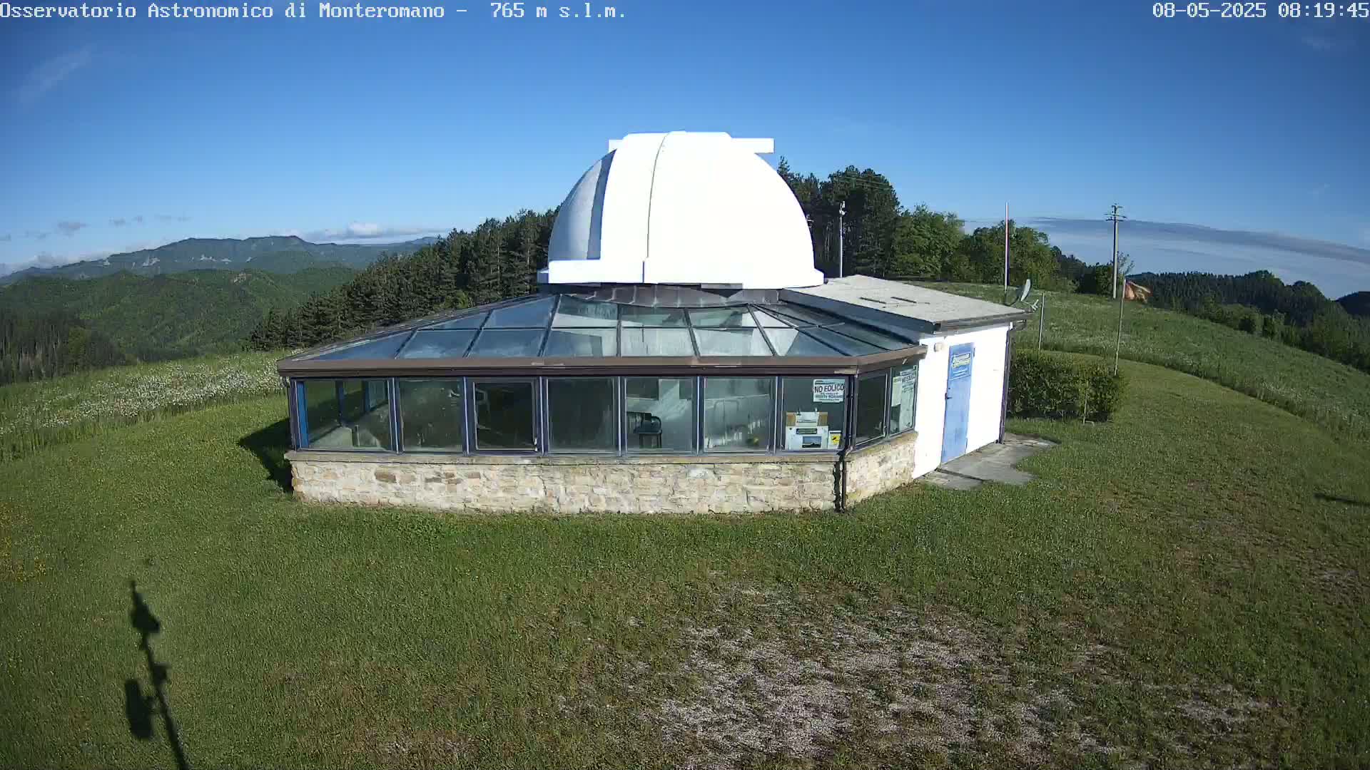 A stone building with a glass enclosure and a white dome sits on a grassy hilltop under a clear blue sky, with distant mountains visible in the background.