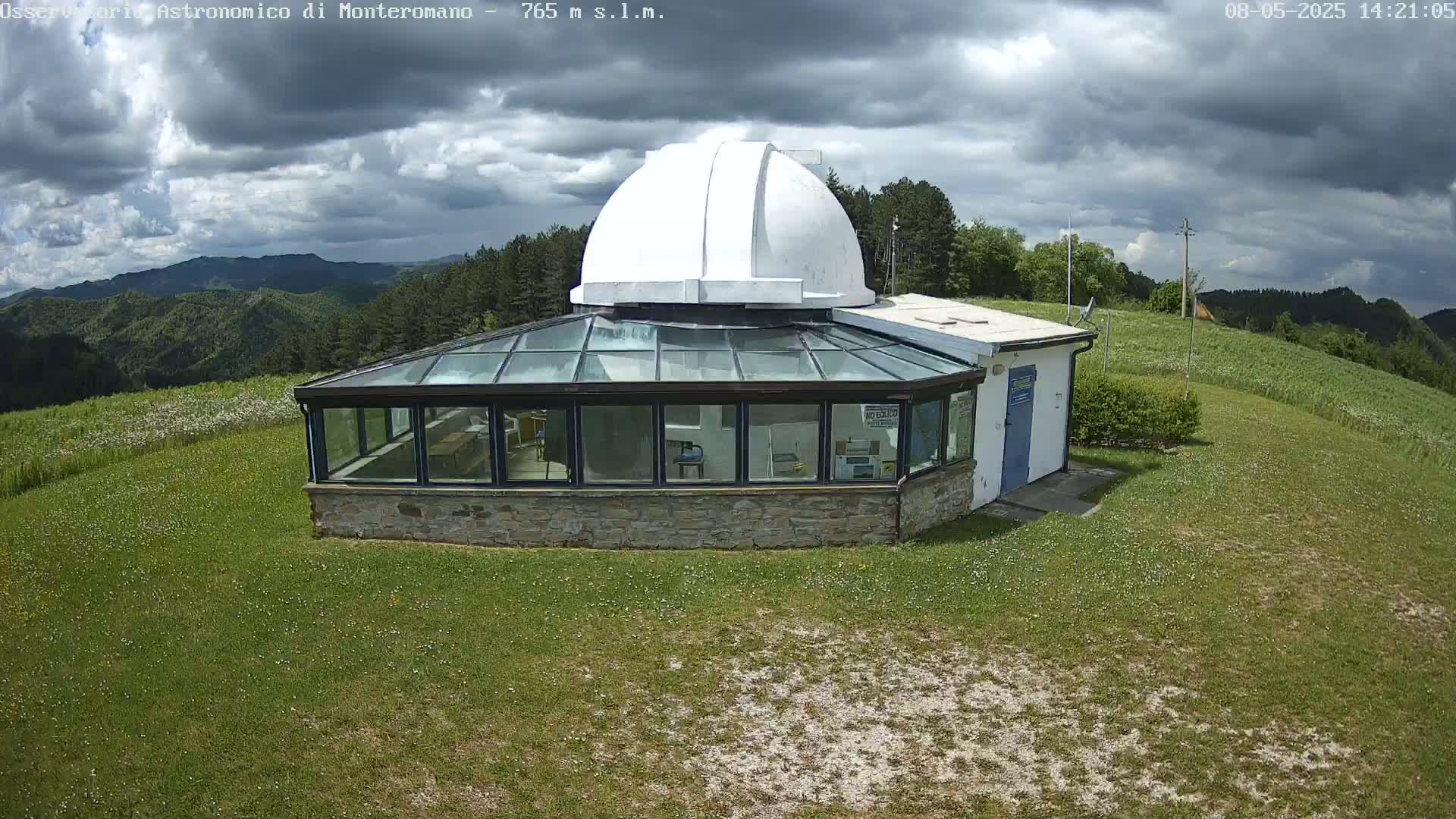 A glass-roofed building with an attached white dome sits on a grassy hill overlooking a mountain range under a cloudy sky.