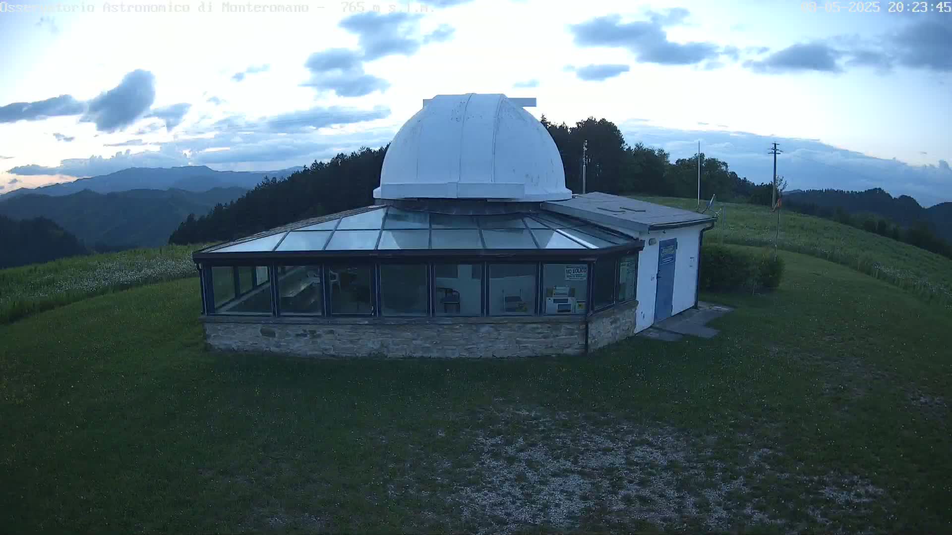 A stone building with a glass roof and a white dome sits on a grassy hilltop under a partly cloudy twilight sky, overlooking a range of mountains.