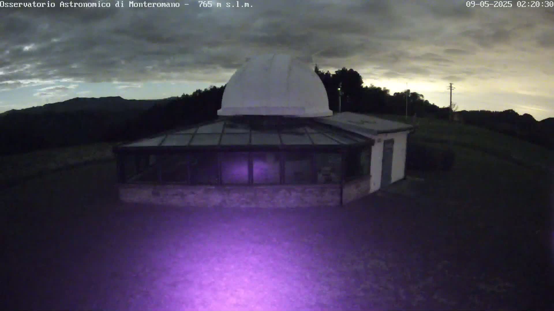 A low-light image shows a white observatory dome atop a building with a glass sunroom, situated on a dark, grassy hill under a cloudy, twilight sky.