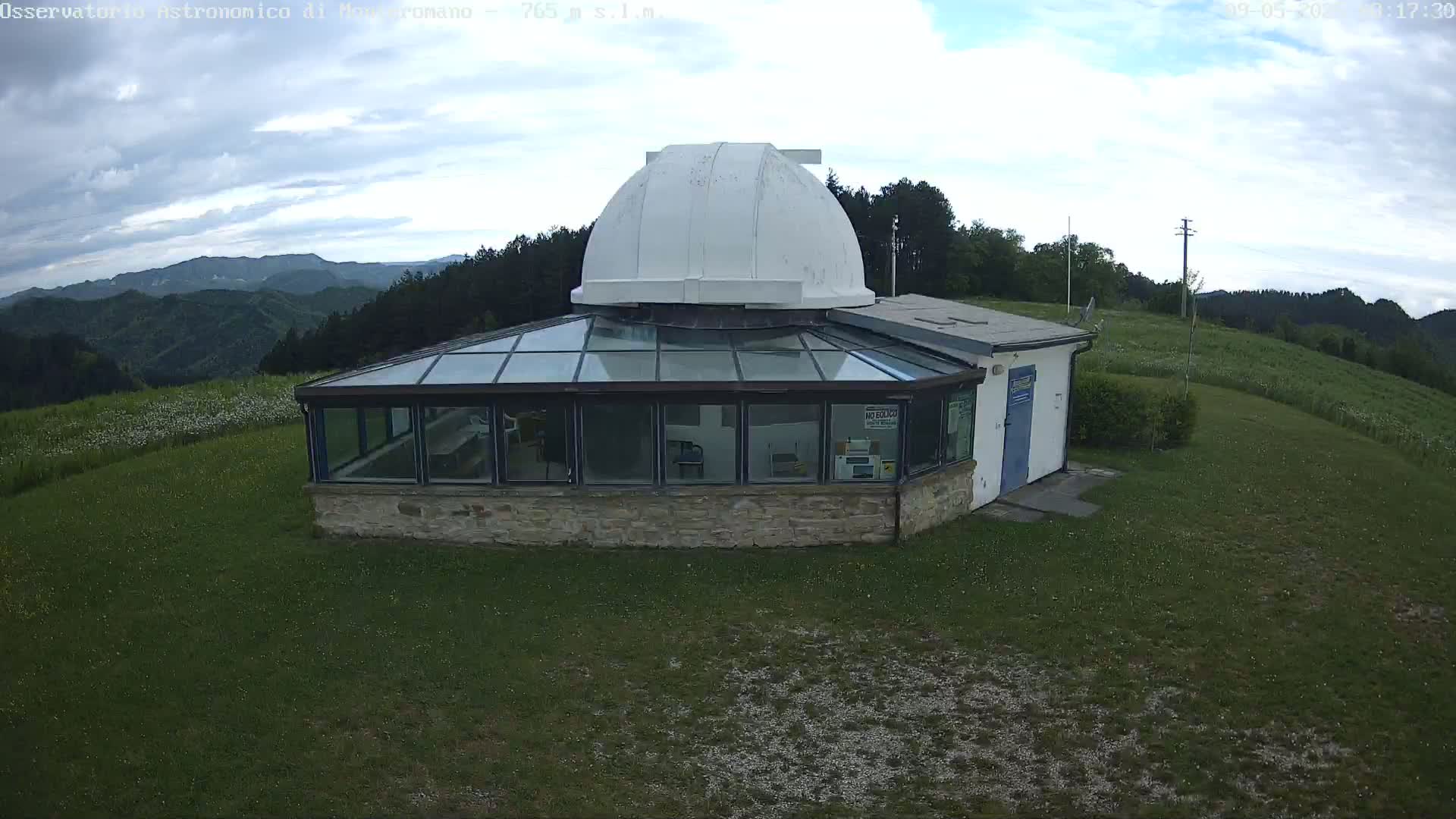 A stone building with a glass-roofed addition and a white dome sits on a grassy hilltop under a partly cloudy sky, overlooking a mountainous landscape.