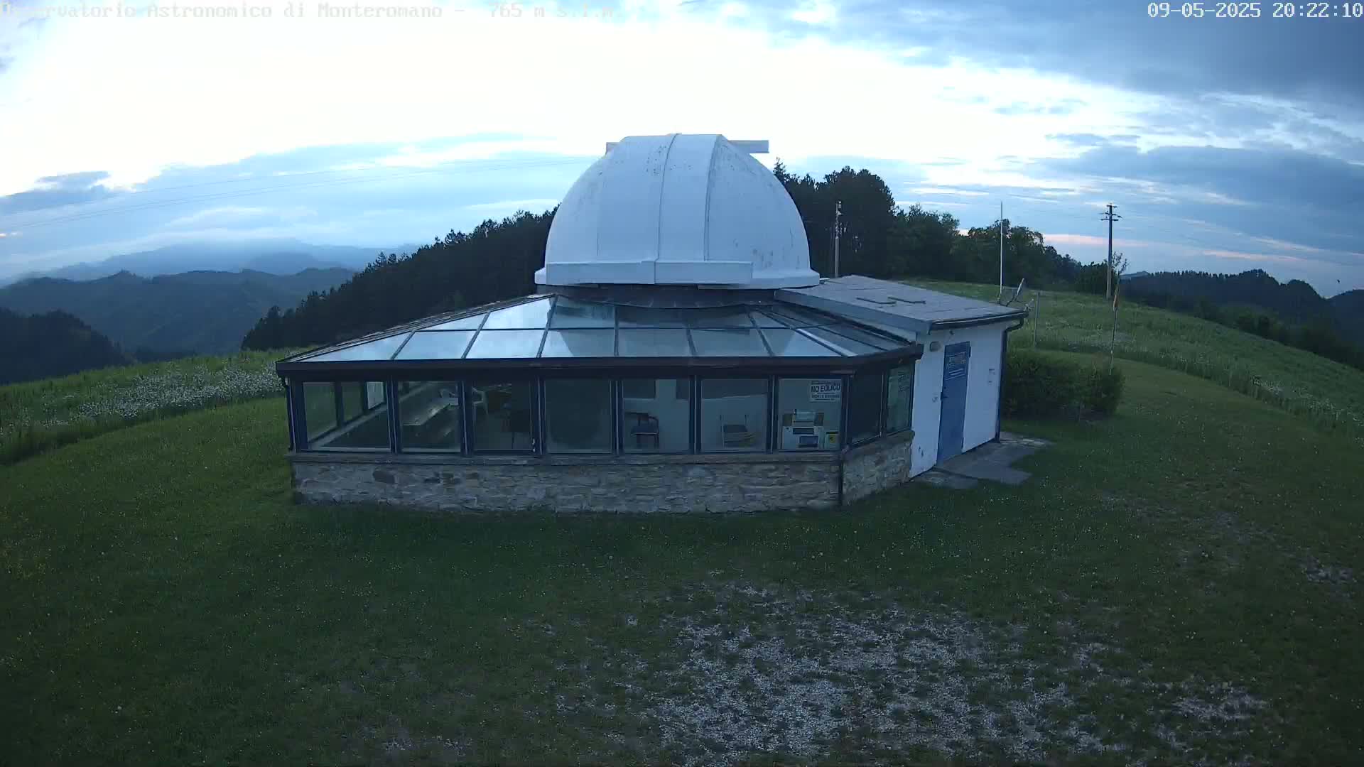 A stone building with a glass sunroom and a white dome on top sits on a grassy hill overlooking a range of mountains under a partly cloudy twilight sky.