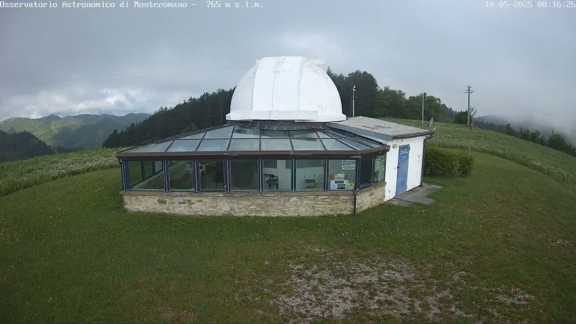 A stone building with a glass sunroom and a white dome sits on a grassy hill overlooking a hazy mountain range under an overcast sky.