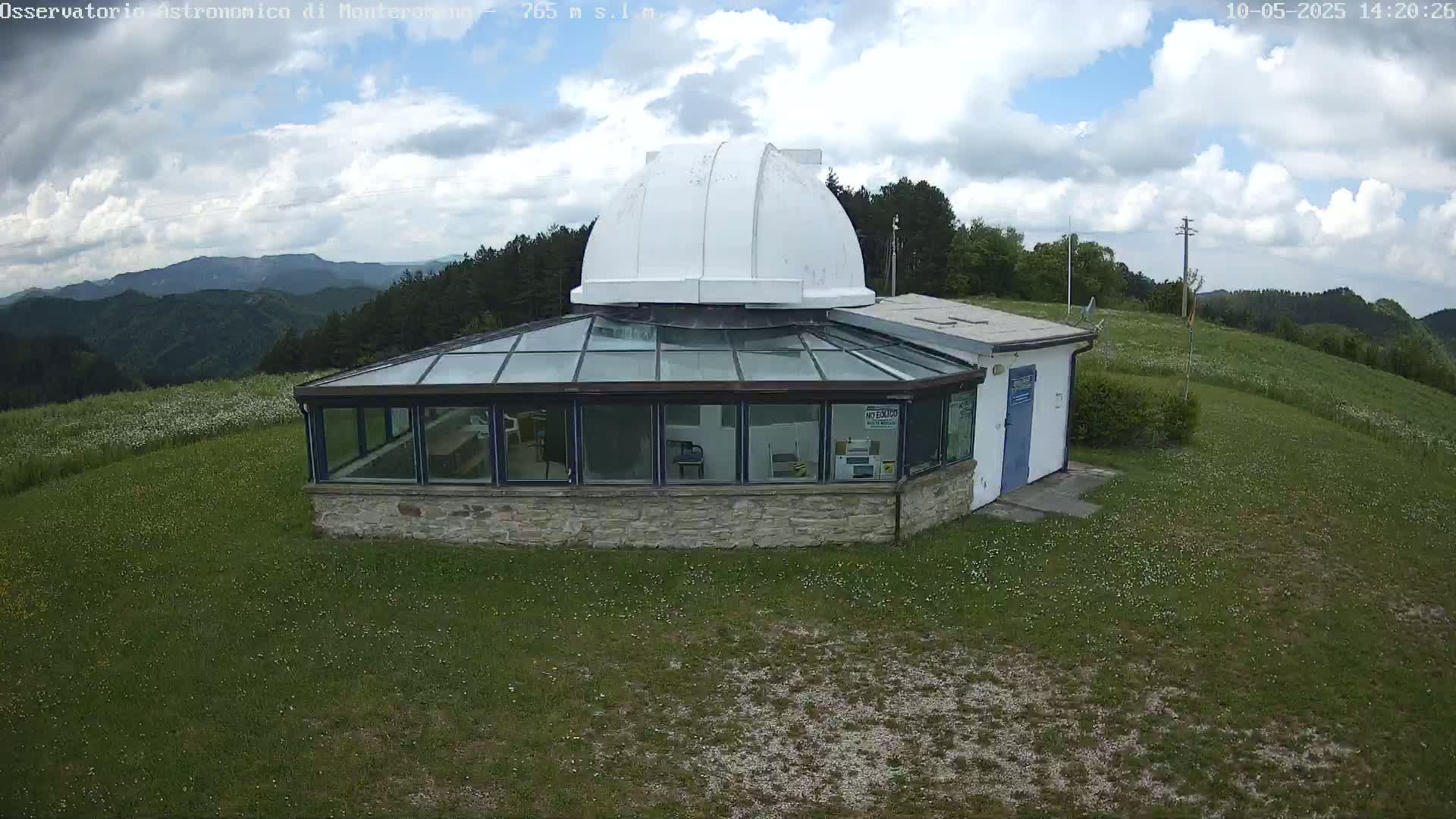 A white domed observatory building with a glass-roofed addition sits on a grassy hilltop under a partly cloudy sky.