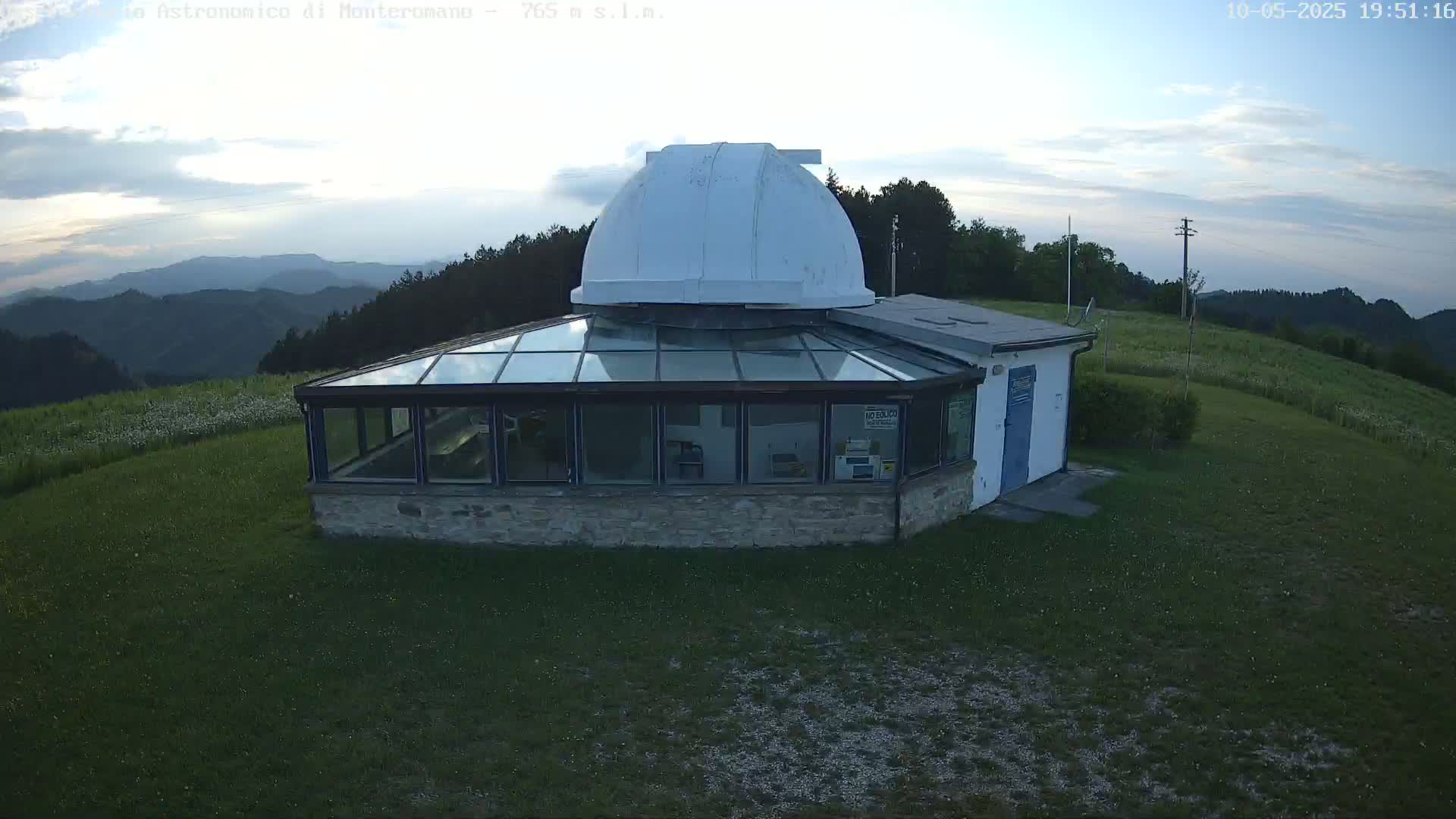A stone building with a glass roof and a white dome sits on a grassy hilltop overlooking a range of mountains under a partly cloudy evening sky.