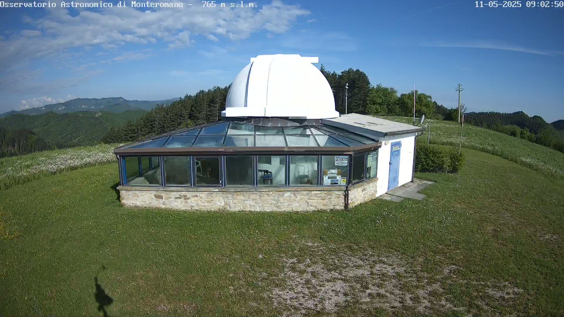 A white dome observatory building with a glass-roofed addition sits on a grassy hilltop under a mostly clear blue sky.
