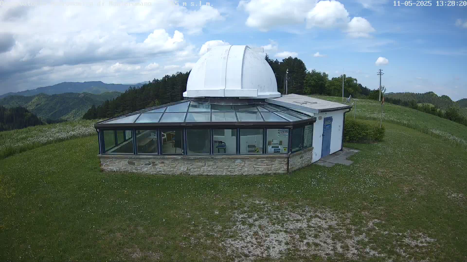 A stone building with a glass roof and a white dome sits on a grassy hilltop under a partly cloudy sky, overlooking a range of forested mountains.