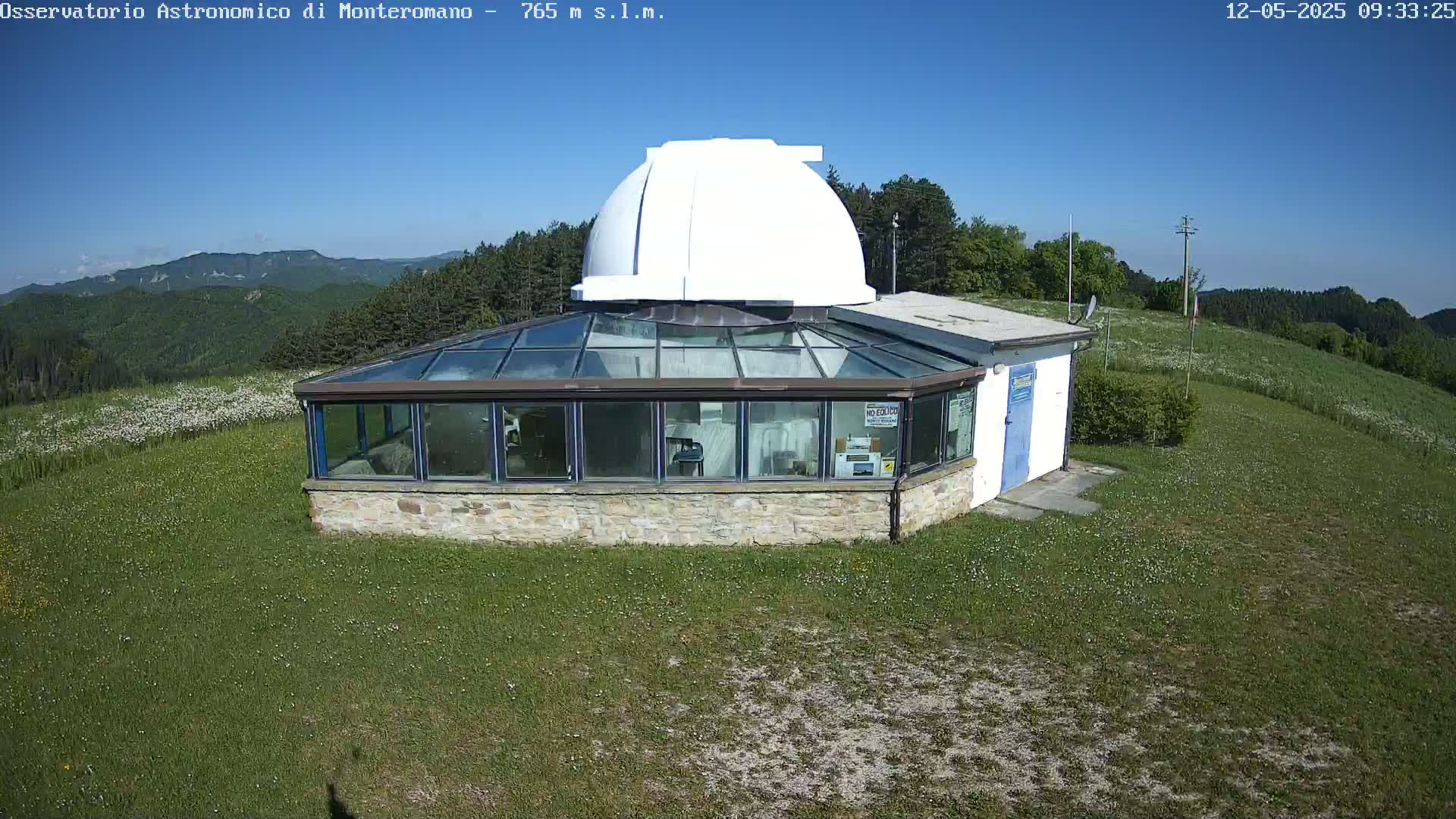 A white-domed observatory building with a glass sunroom sits on a grassy hill overlooking a mountain range under a clear blue sky.