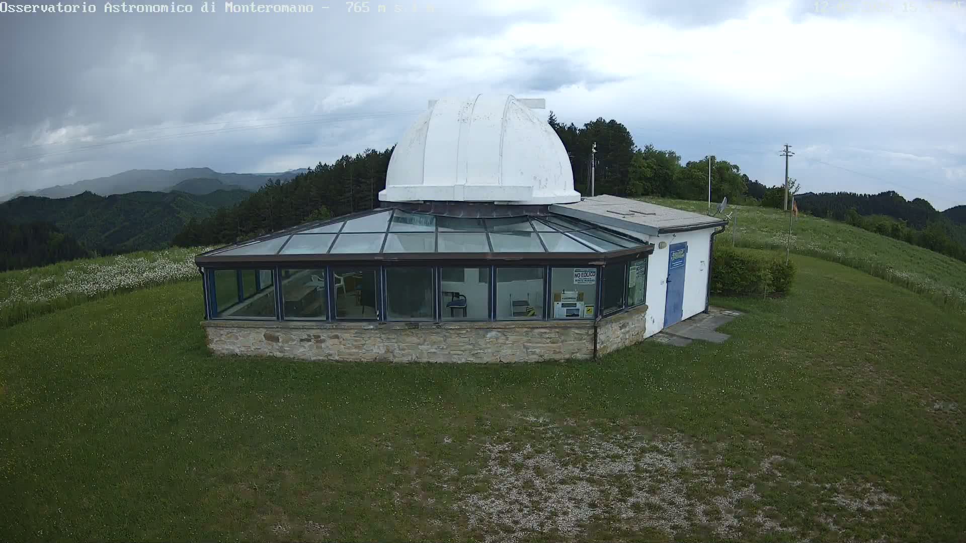A stone building with a glass-roofed addition and a white dome sits on a grassy hilltop under a cloudy sky, overlooking a mountainous landscape.