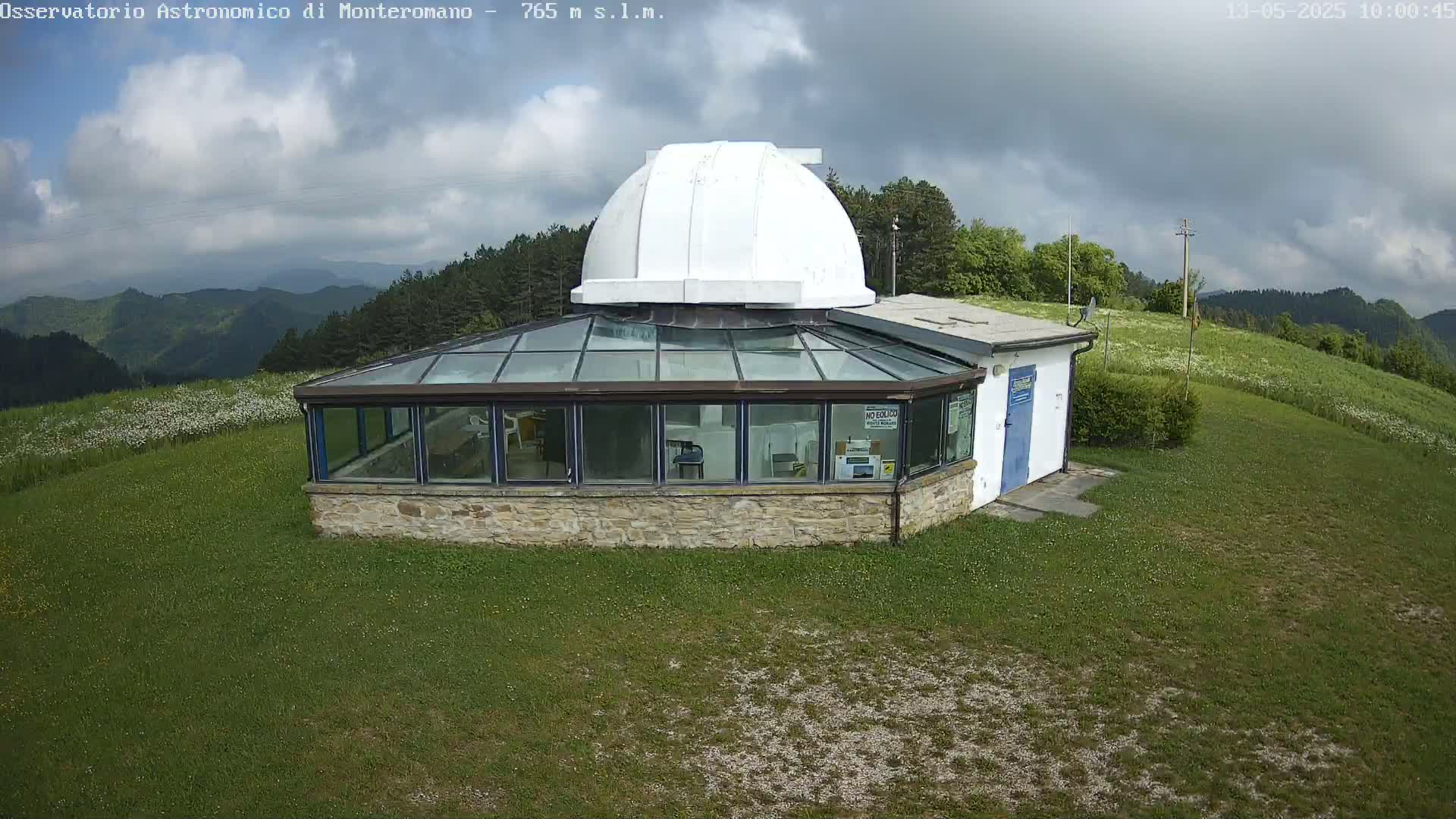 A stone building with a glass roof and a white dome sits on a grassy hill under a partly cloudy sky.