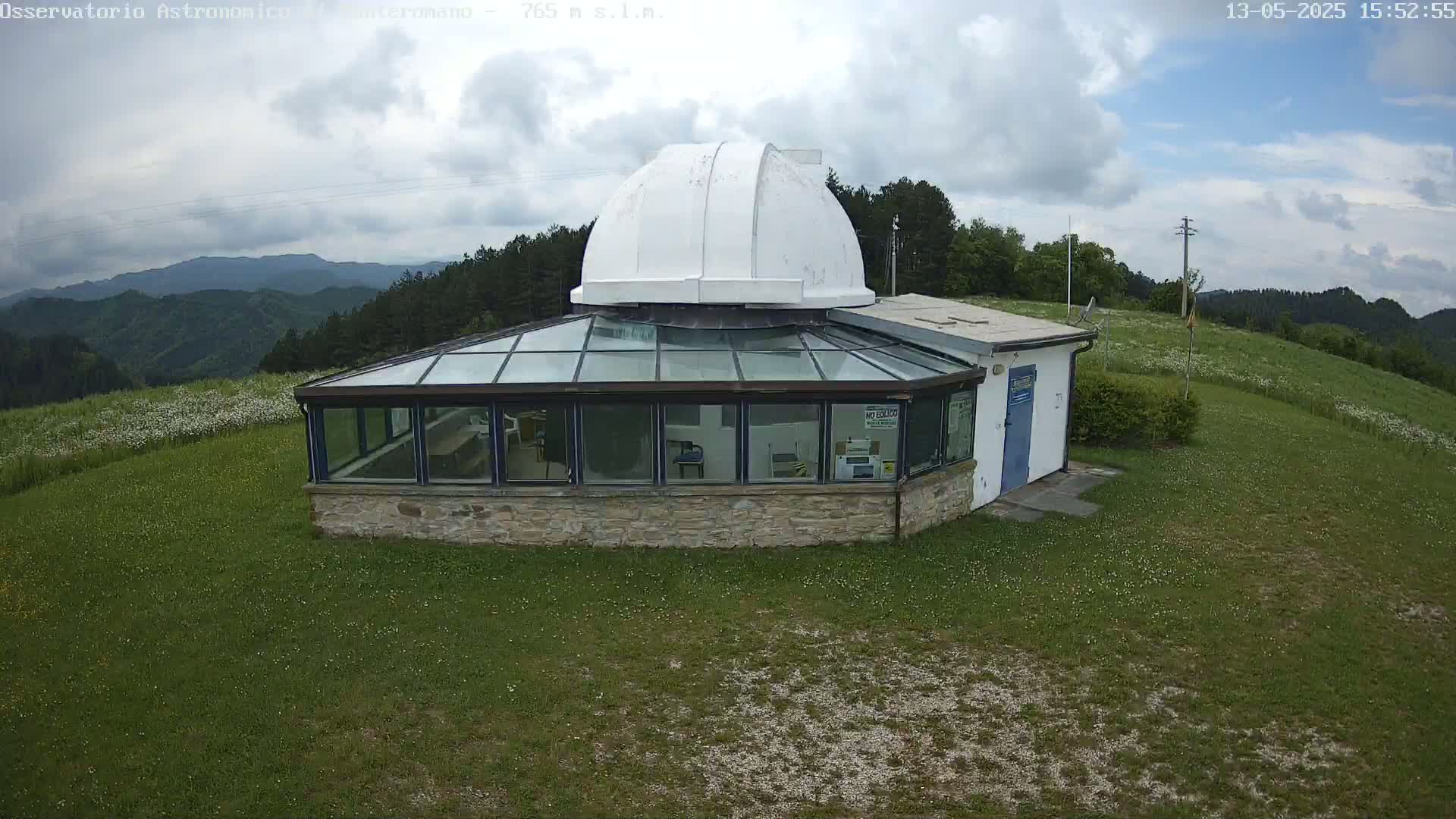 A white domed observatory building with a glass sunroom is situated on a grassy hill overlooking a mountainous landscape under partly cloudy skies.