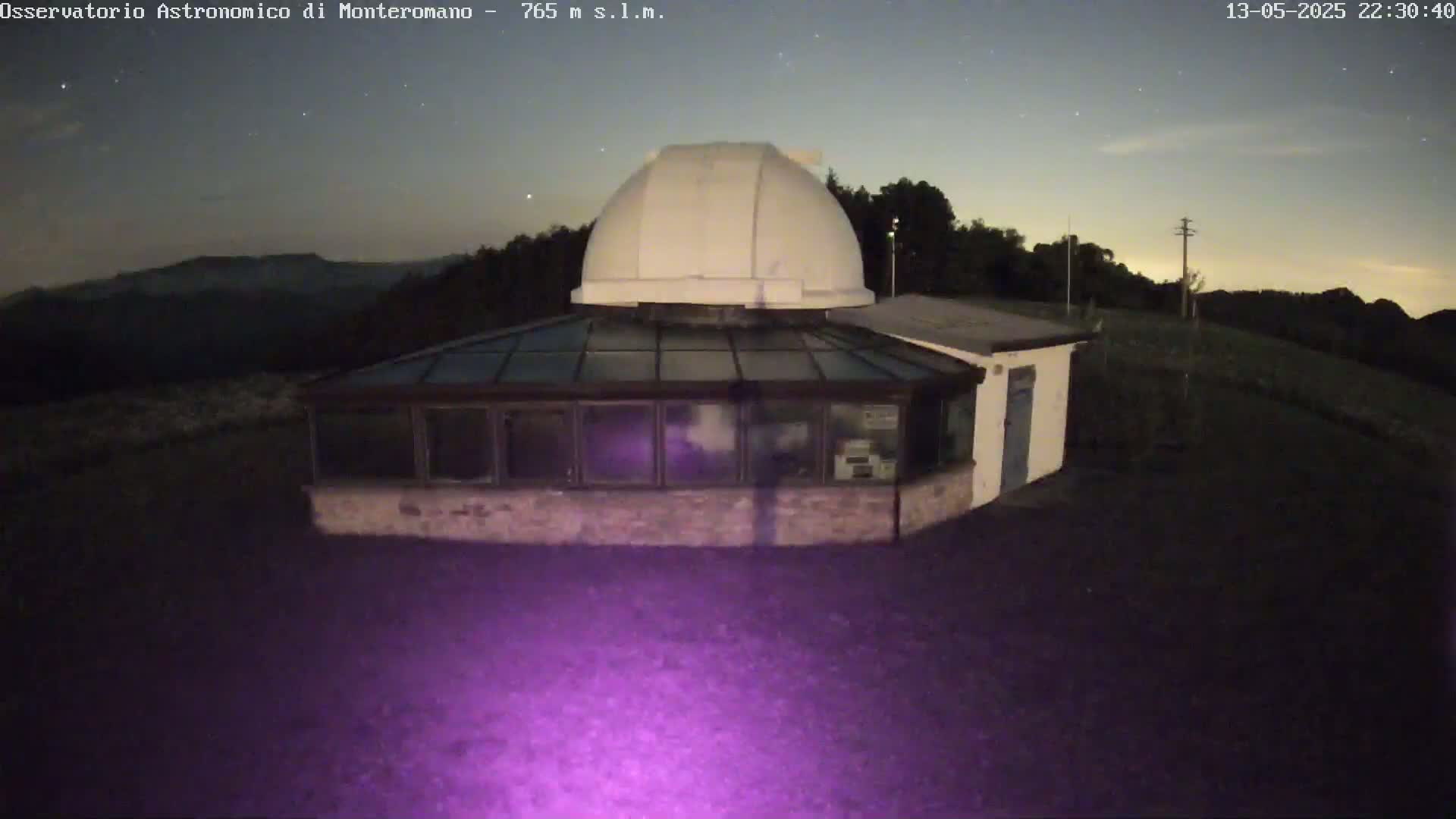 A low-light, nighttime image shows a stone building with a glass-roofed addition and a white dome on top, situated in a dark, mostly flat area with distant hills under a mostly clear, starry sky.