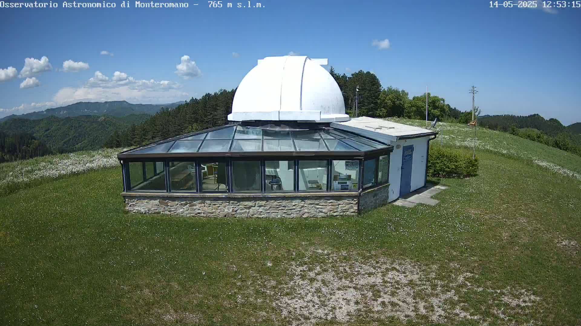 A stone building with a glass sunroom and a white dome sits on a grassy hilltop under a mostly sunny sky, overlooking a green, hilly landscape.