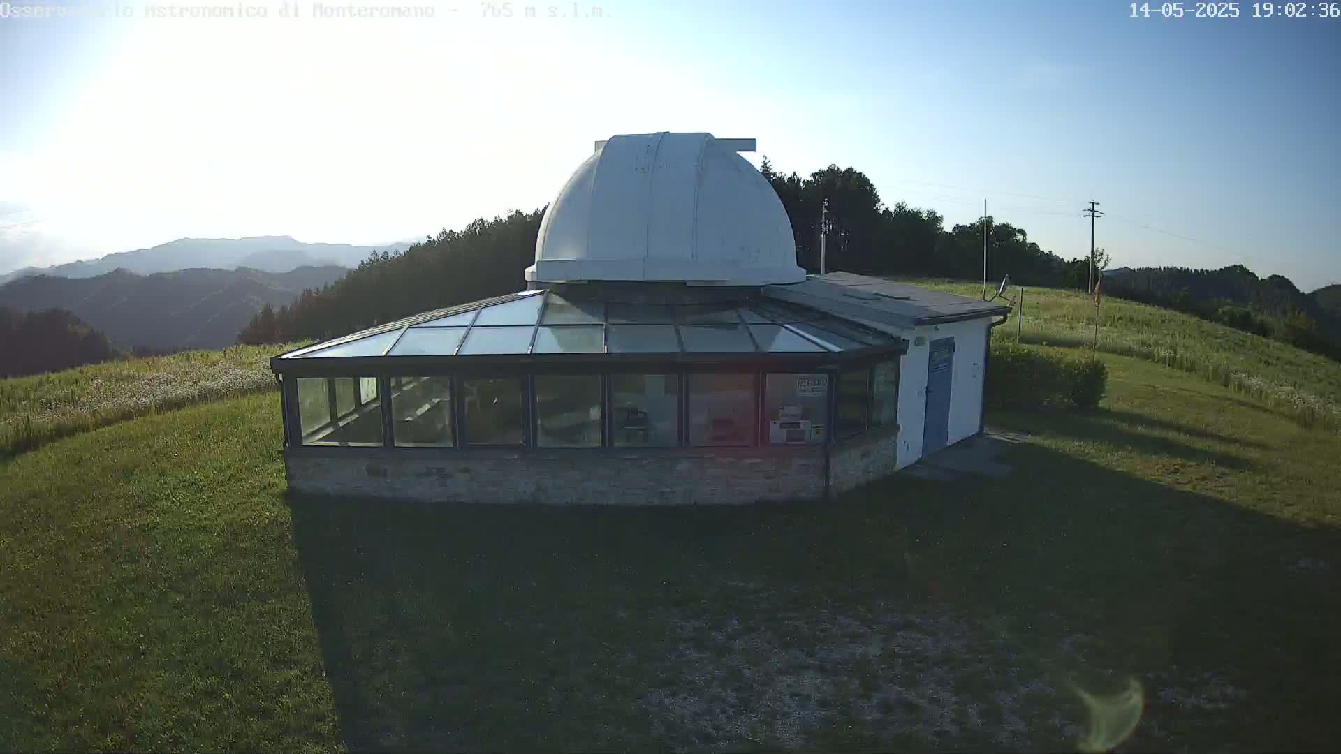 A white dome observatory sits atop a stone building with a glass-roofed addition, overlooking a grassy field and distant mountains under a clear, bright sky.