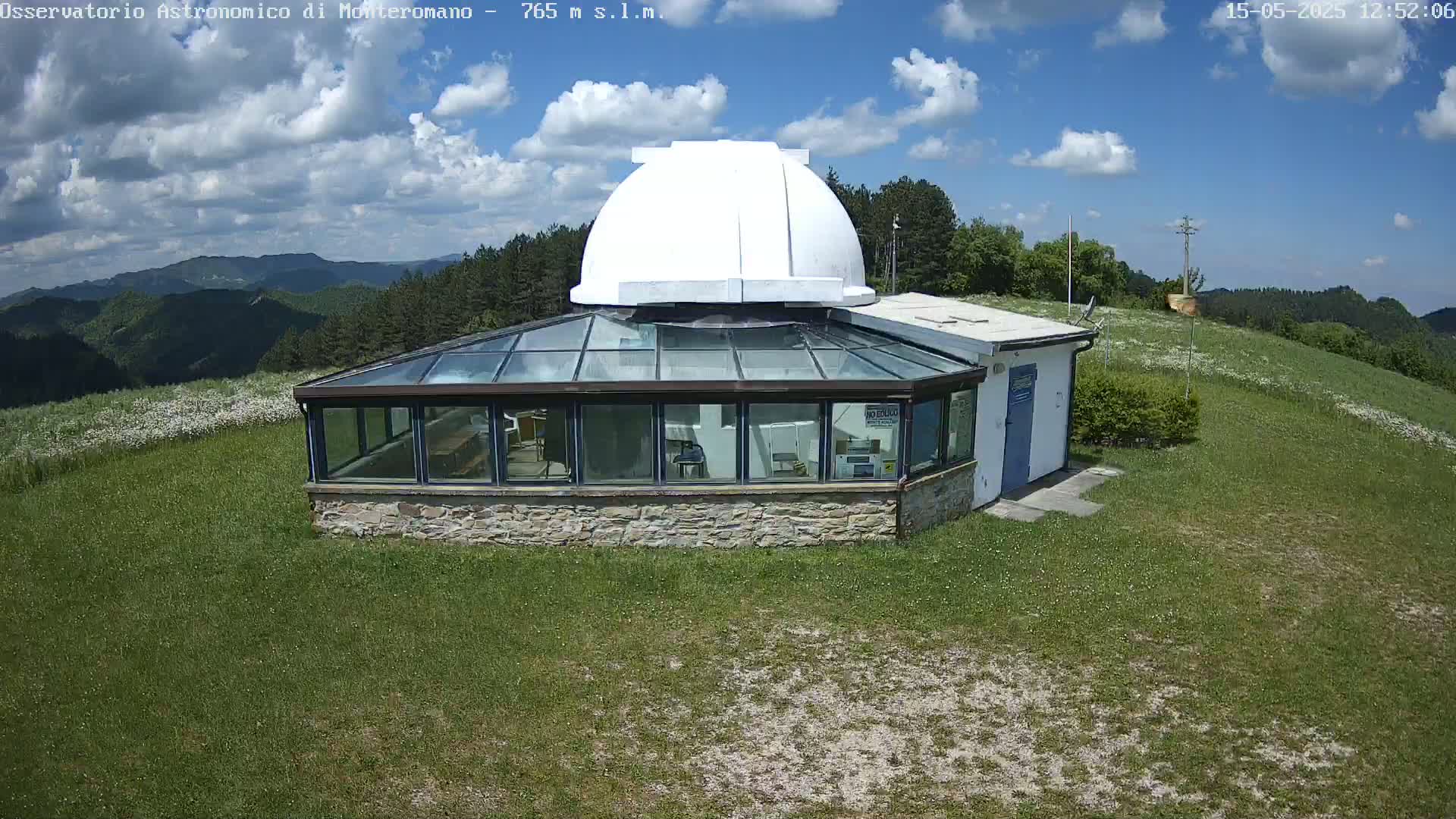 A stone observatory building with a glass enclosure and a white dome sits on a grassy hill under a partly cloudy blue sky.