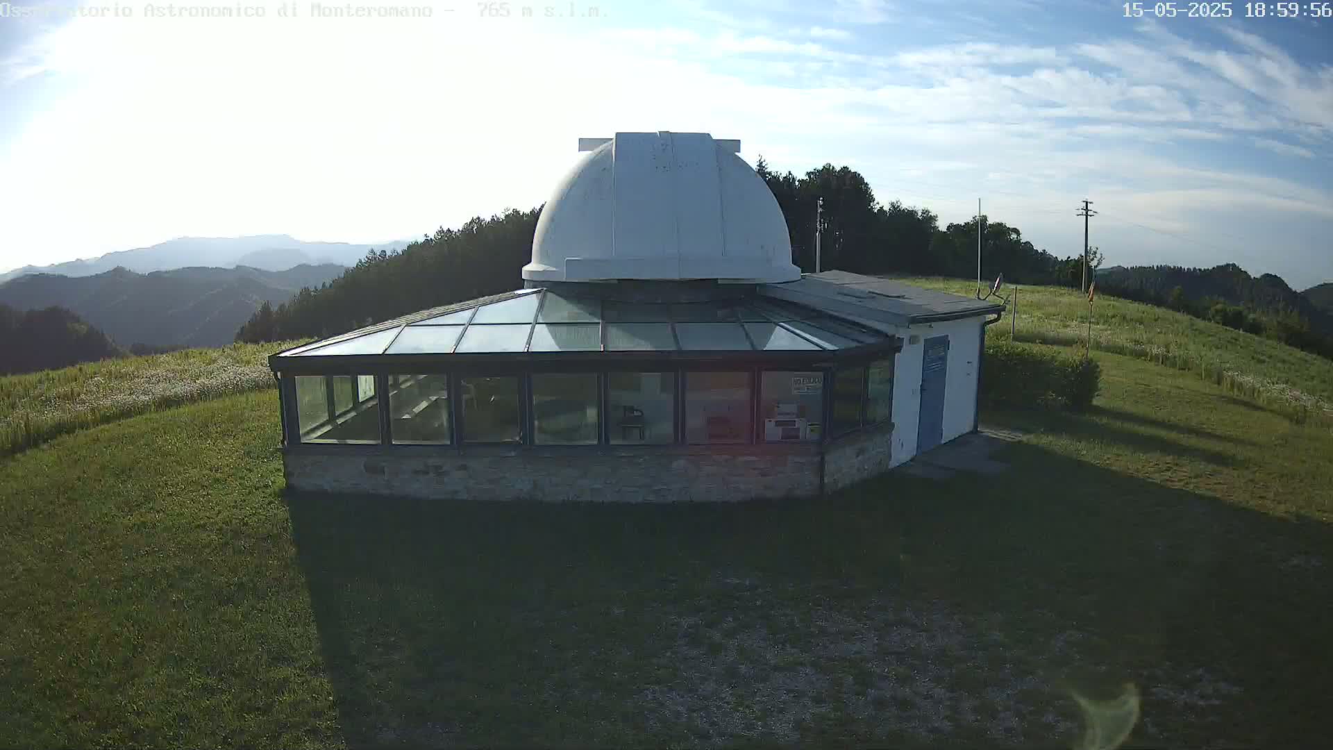 An observatory with a glass-roofed addition sits on a grassy hilltop under a mostly clear, sunny sky, overlooking a mountain range.
