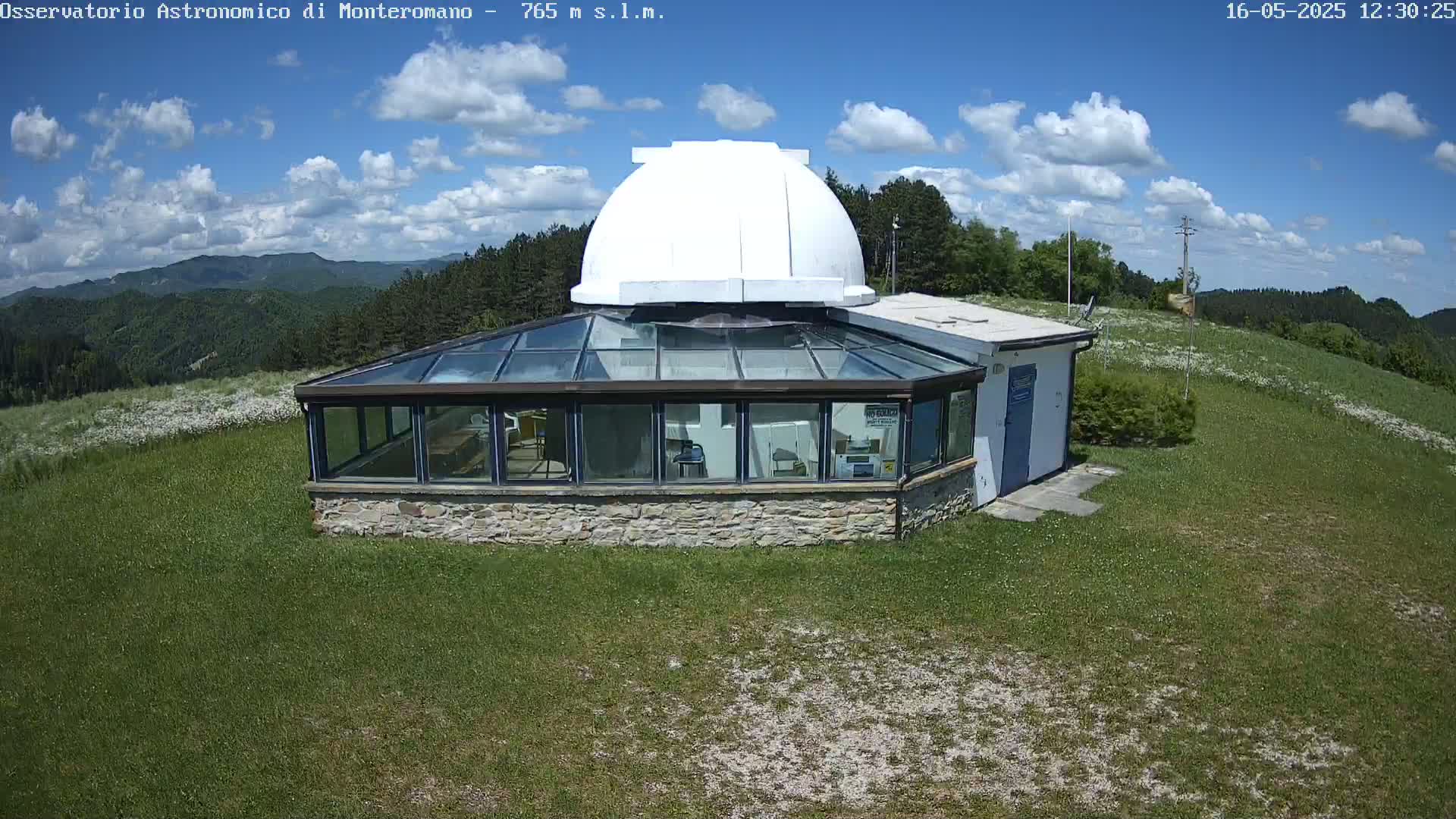 A stone building with a glass enclosure and a white dome sits on a grassy hilltop under a partly cloudy blue sky.