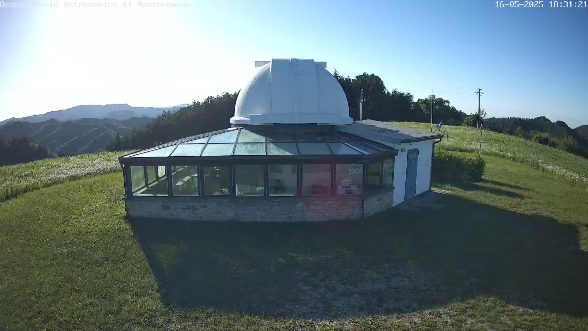 A white observatory dome sits atop a stone building with a glass sunroom, overlooking a grassy hillside and distant mountains under a clear, sunny sky.