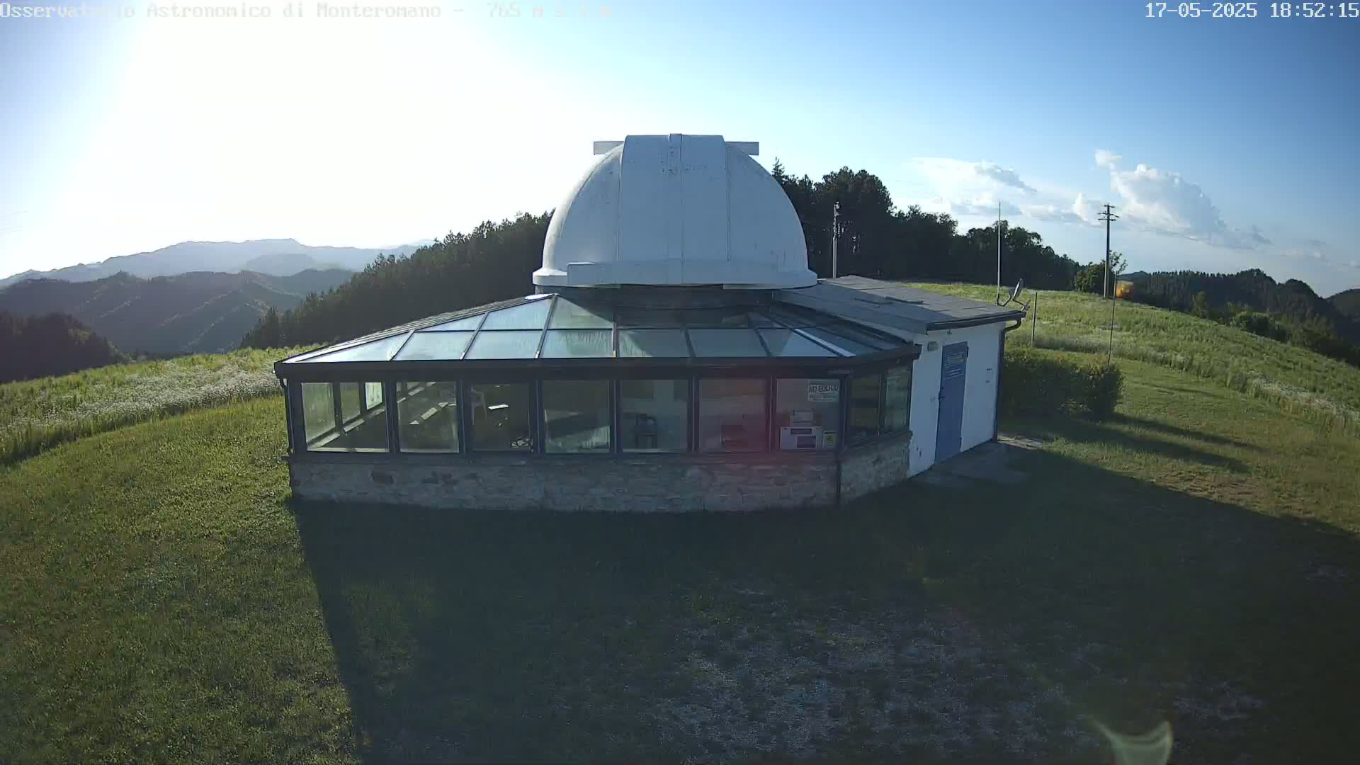 A white-domed observatory building with a glass sunroom sits on a grassy hilltop under a mostly clear, sunny sky, overlooking a mountainous landscape.