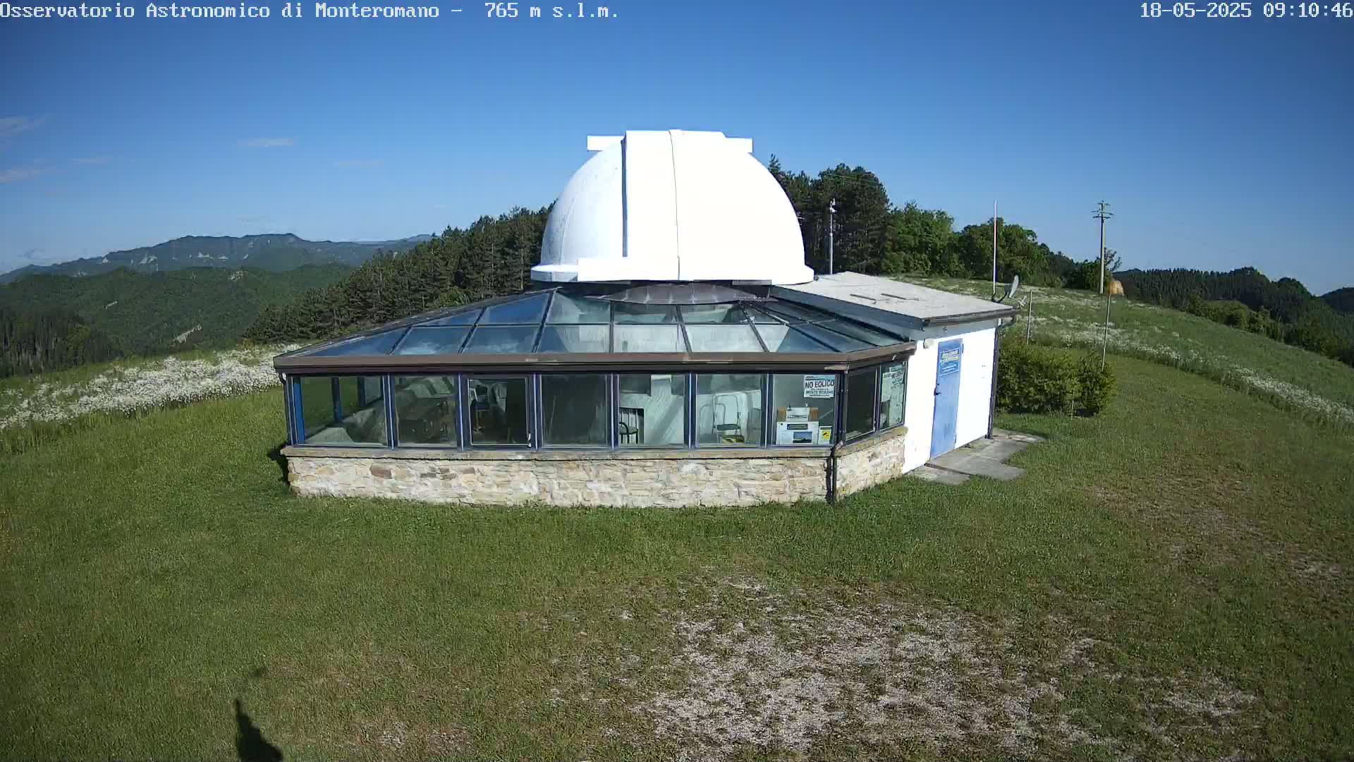 A white dome observatory sits atop a grassy hill on a clear, sunny day, overlooking a mountain range in the distance.