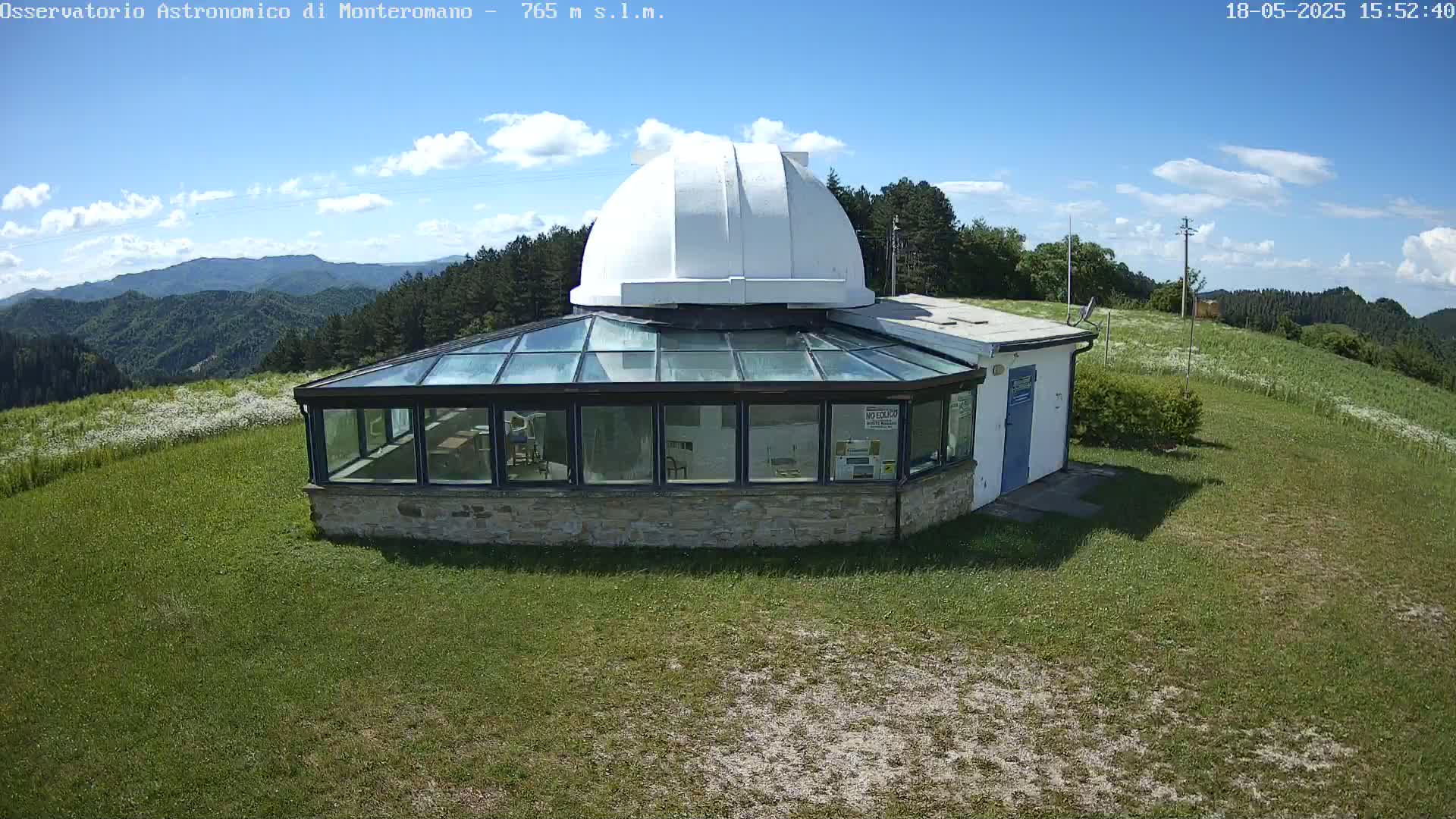 A white dome-shaped observatory building with a glass sunroom sits on a grassy hilltop overlooking a mountainous landscape under a mostly sunny sky.