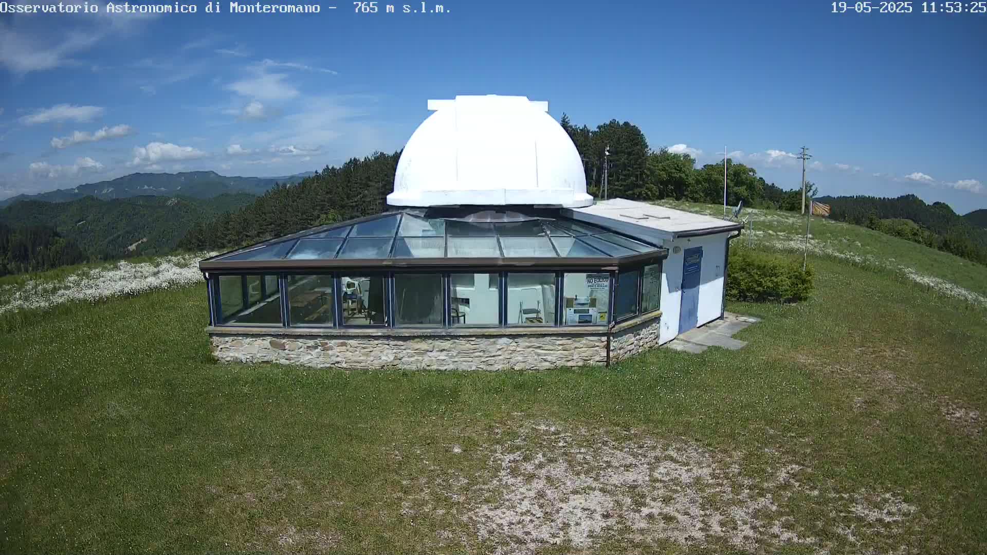 A stone building with a glass roof and a white dome sits on a grassy hilltop under a mostly sunny sky.