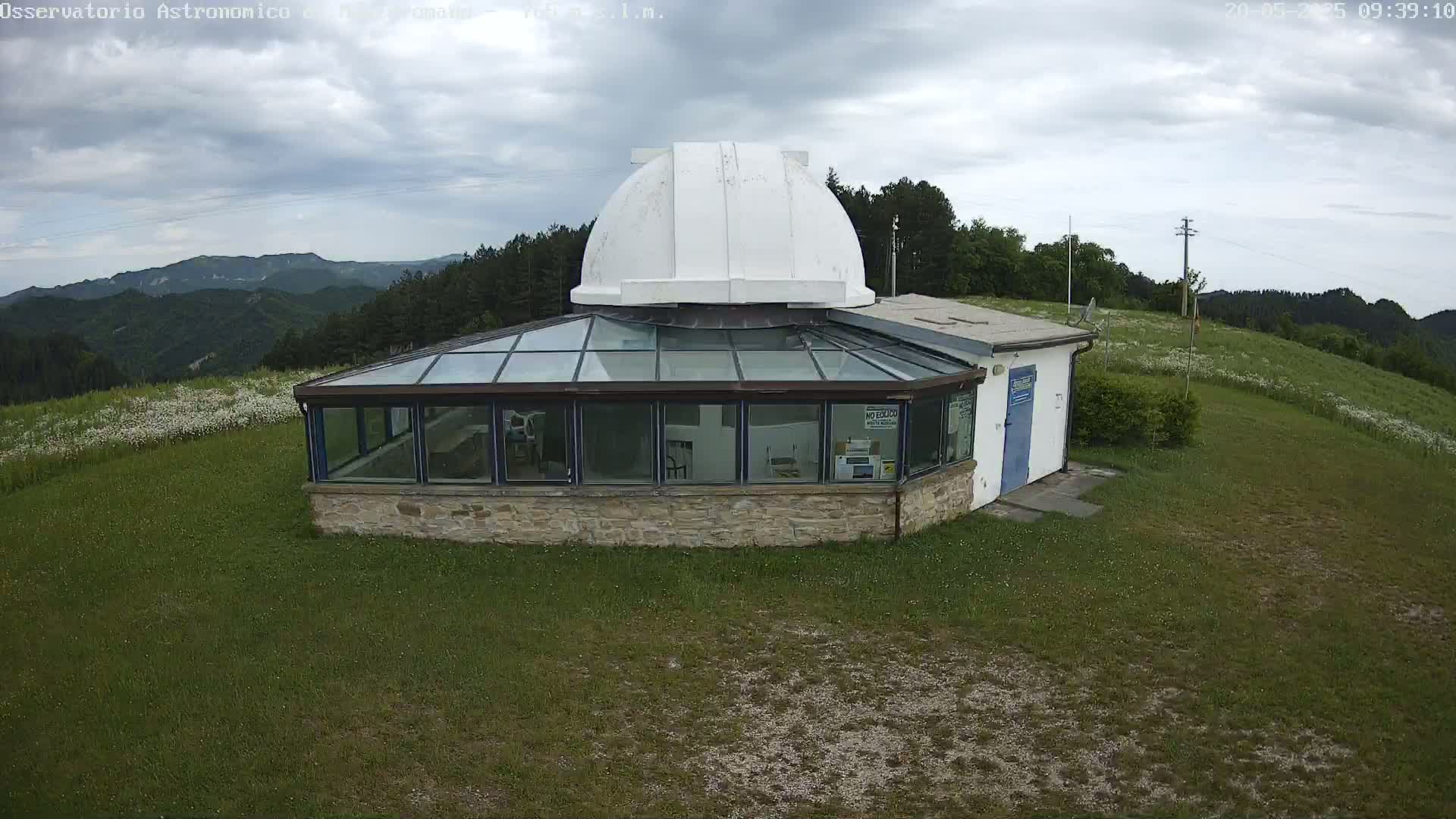An observatory with a white dome sits atop a grassy hill under a cloudy sky, overlooking a mountainous landscape.